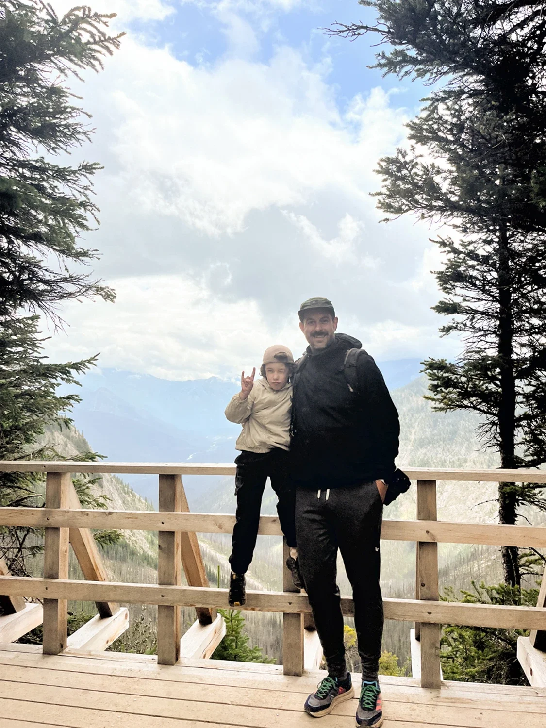 A man and a young boy standing on a wooden platform in a forested mountain area. The boy is making a peace sign with his fingers, and both are smiling. Mountains and cloudy sky are visible in the background.