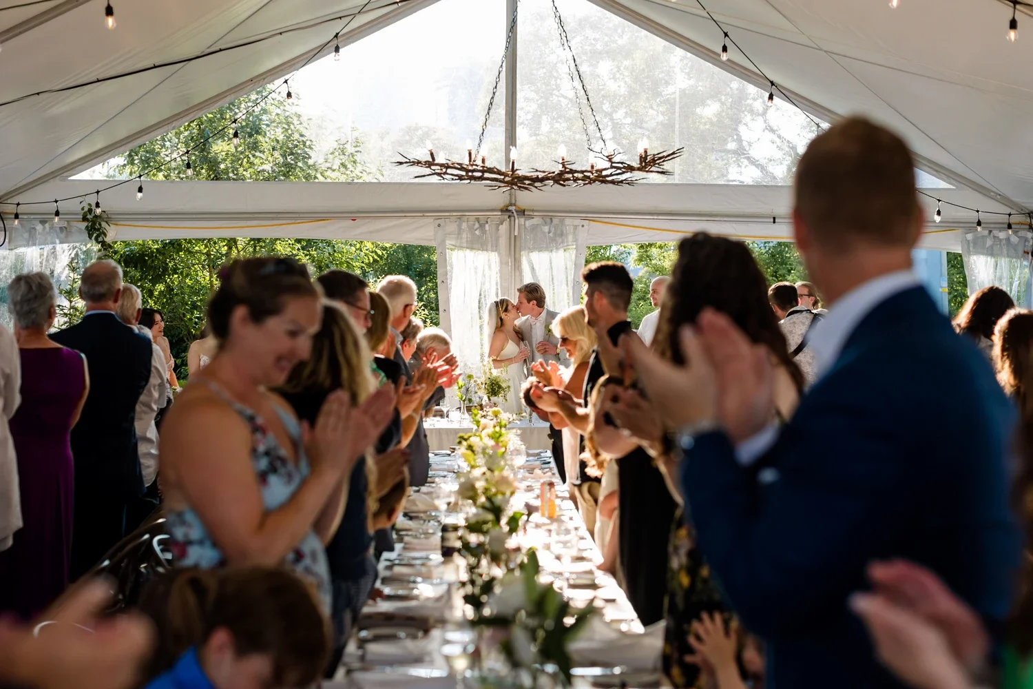 People holding hands and clapping at a wedding reception under a large white tent with string lights and natural sunlight.