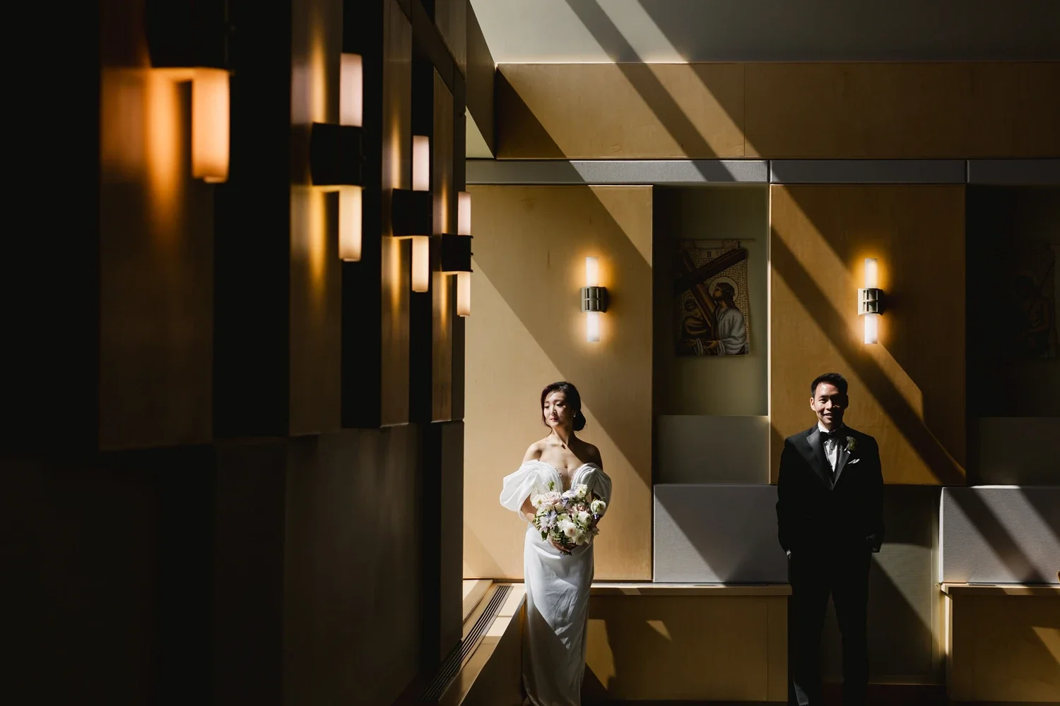 A bride in a white gown holding a bouquet and a groom in a black suit with a bow tie, standing apart in a dimly lit room with modern wall sconces and artwork.