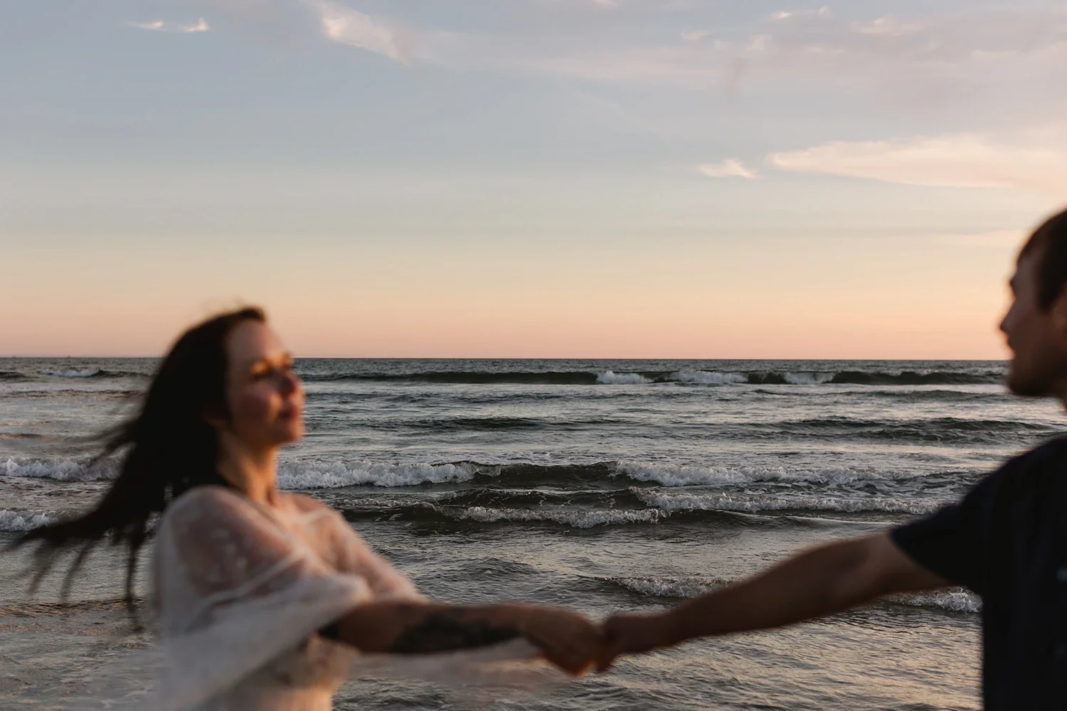 A woman and a man holding hands on the beach at sunset, facing each other with the ocean and sky in the background.