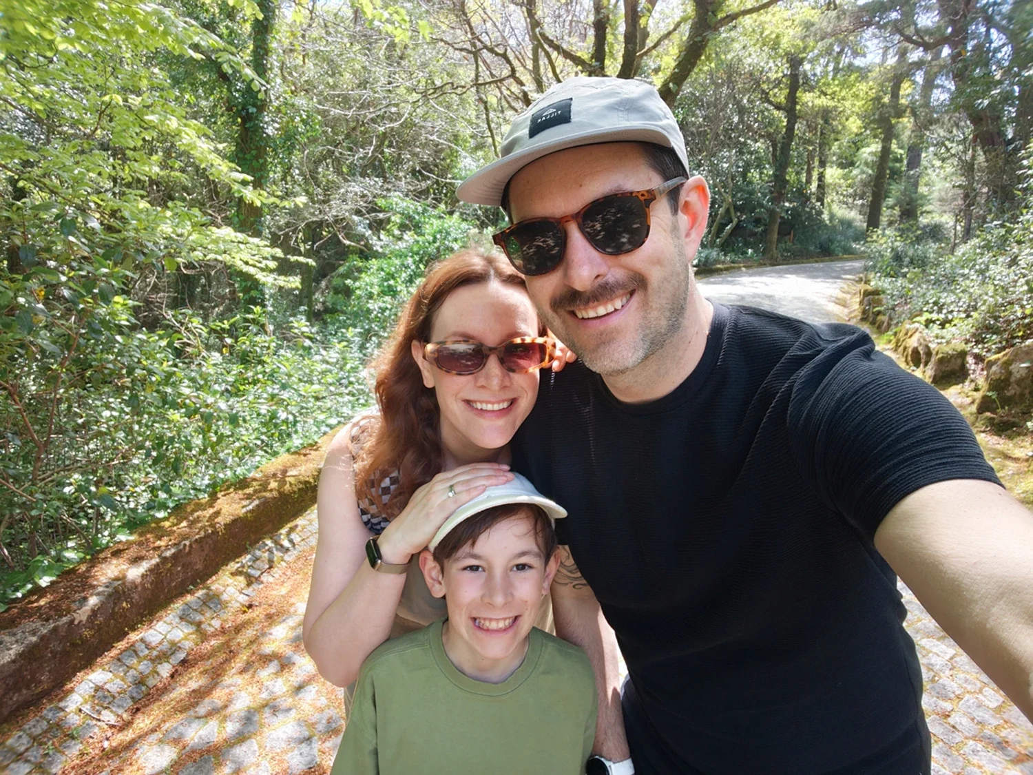 A family of three, with a woman, a man, and a boy, taking a selfie outdoors on a sunny day, surrounded by green trees.
