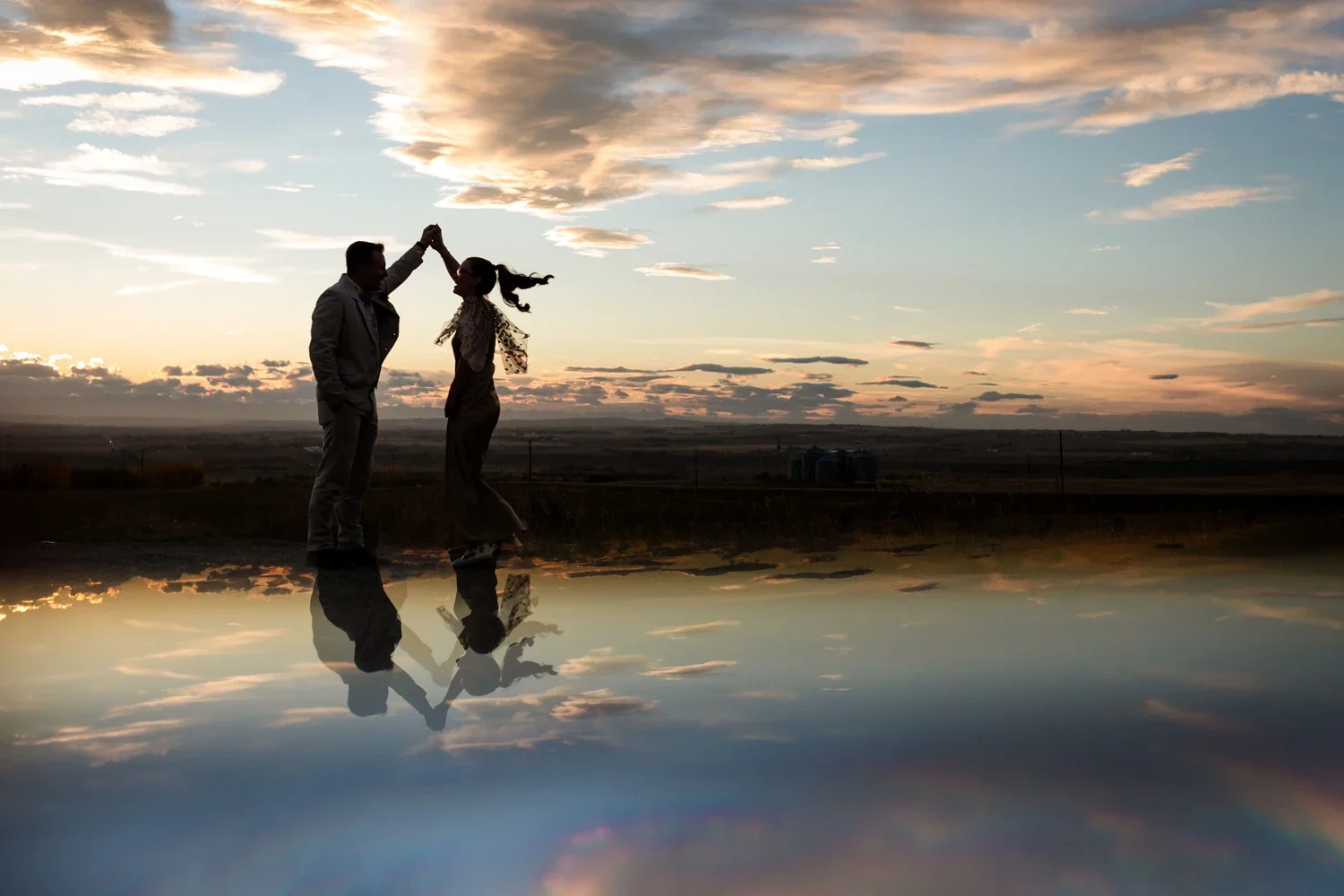 Silhouettes of a couple dancing outdoors during sunset, with clouds in the sky and their reflections visible on a reflective surface in the foreground.