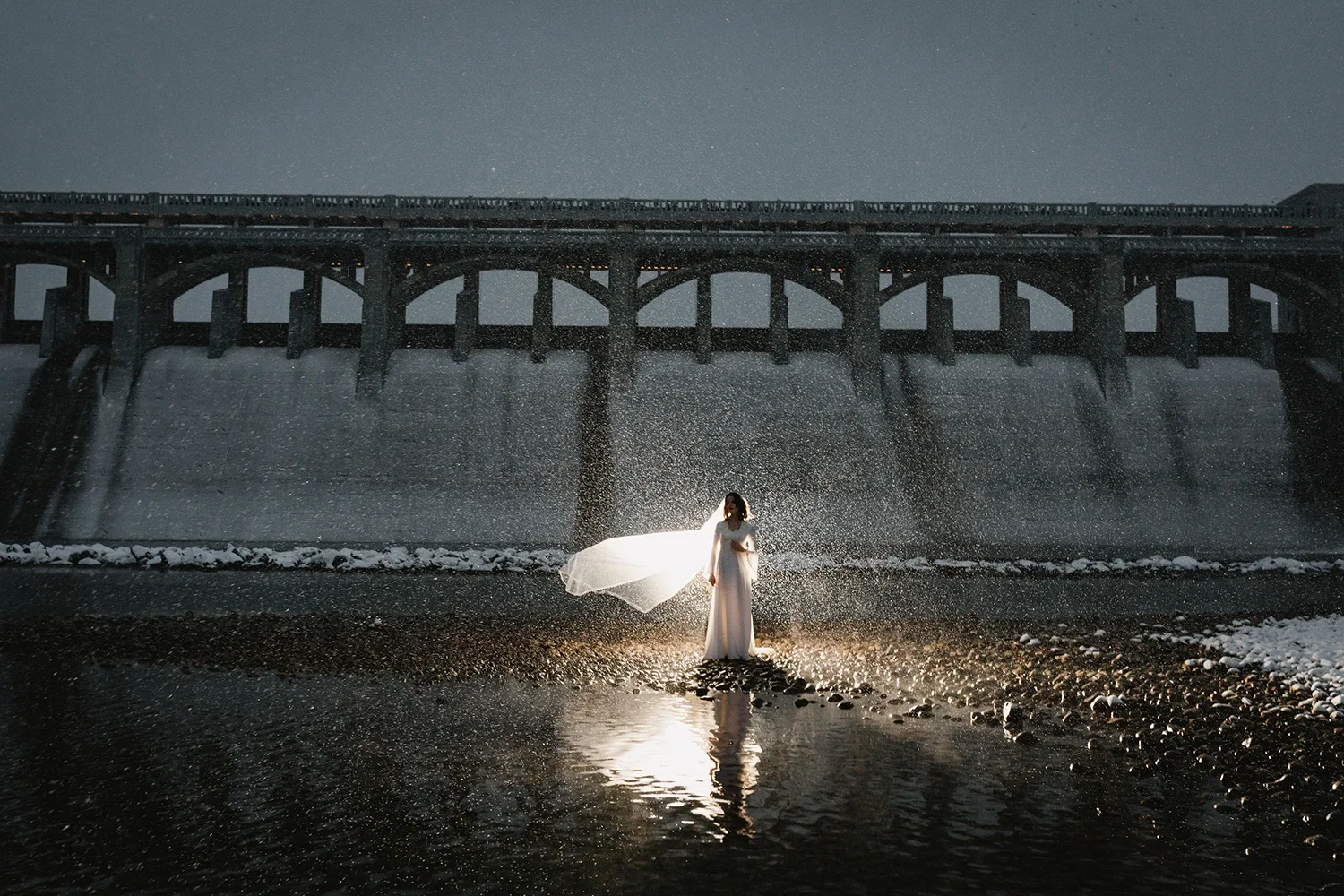 A woman in a white dress and veil stands in water under a large dam at night, illuminated from behind, with rain or spray creating a mist around her and the lights reflecting on the water.