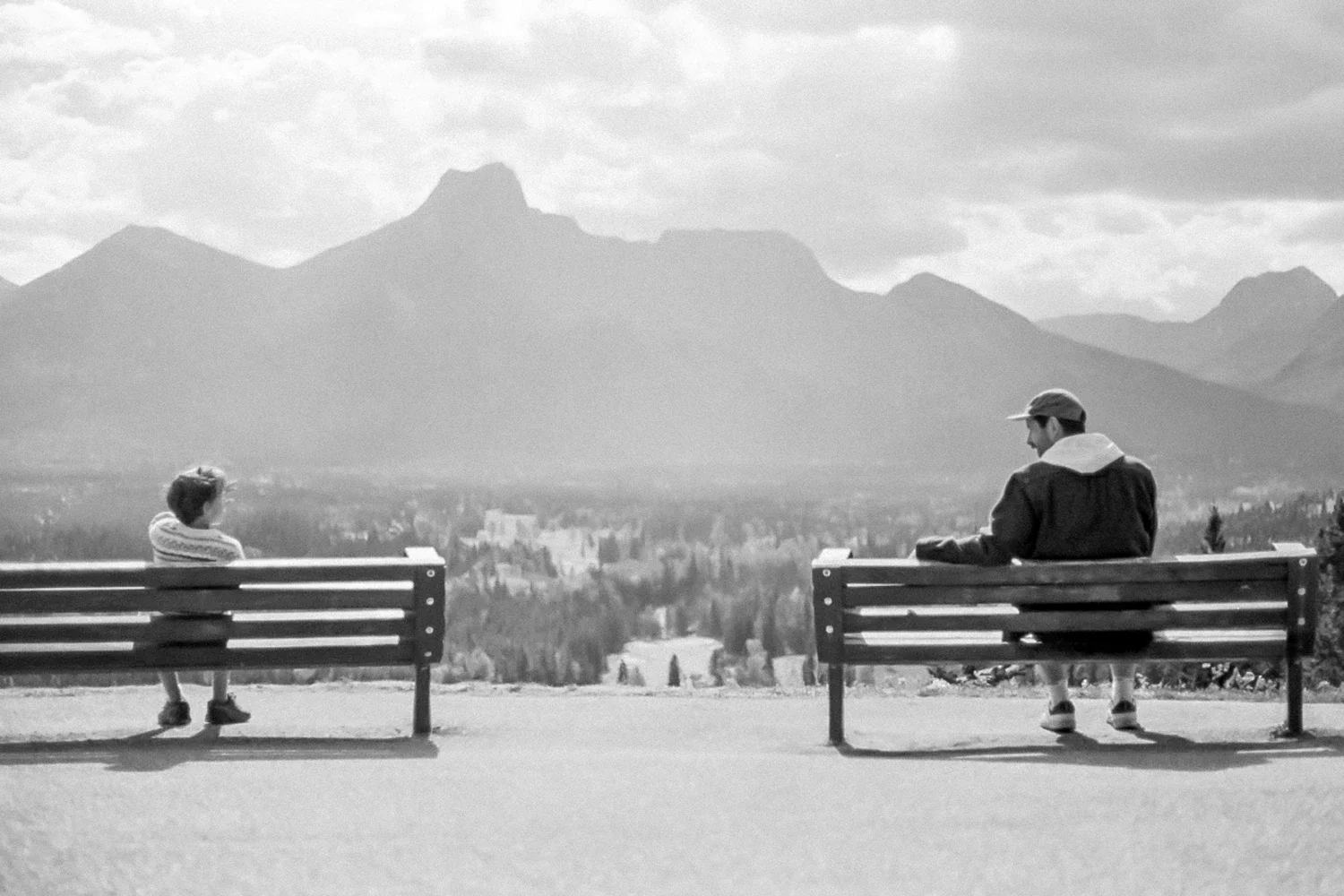 A black and white photo of a woman and a boy sitting on separate benches overlooking a valley with mountains in the background.