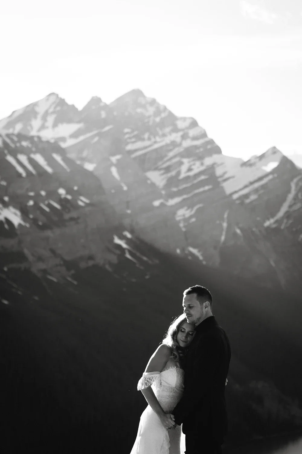 Black and white photo of a couple embracing outdoors with mountain peaks in the background.