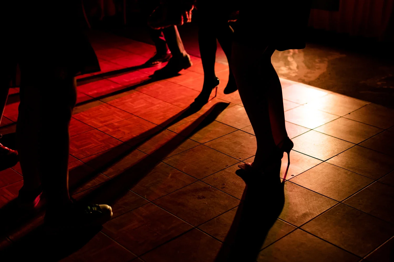 Silhouettes of people dancing in high heels on a dimly lit dance floor with shadows cast on the ground.