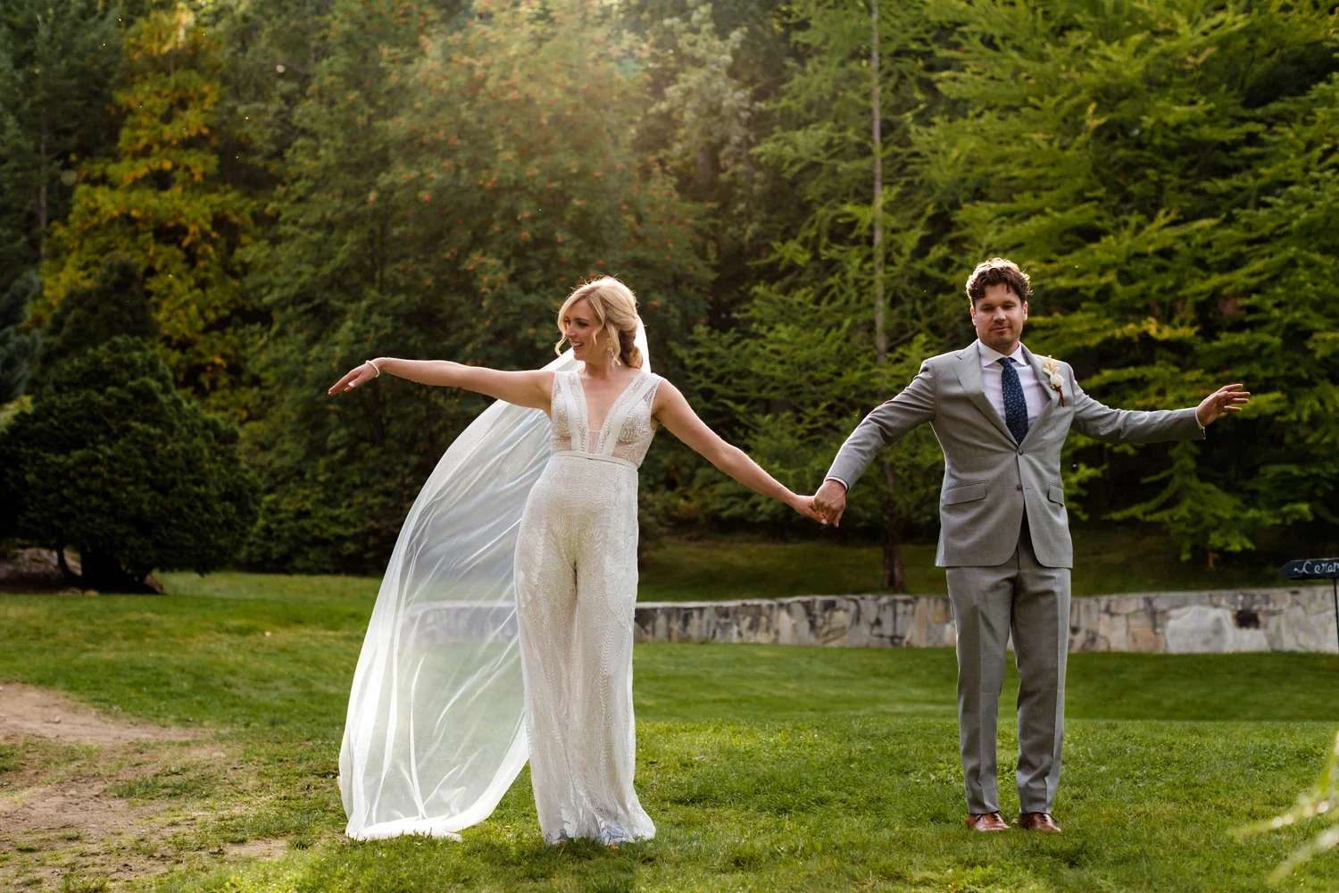 A bride in a white dress holding hands with a groom in a gray suit outdoors during daytime, with green trees in the background.