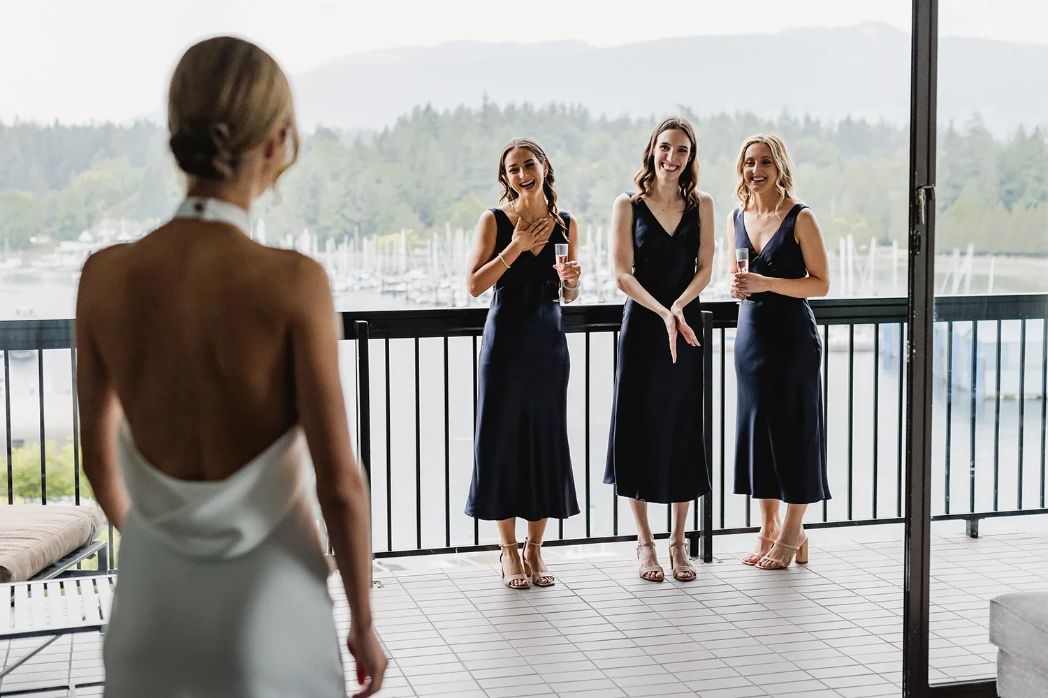 A bride in a white wedding dress standing inside, looking at three bridesmaids outside on a balcony, with a marina and mountains in the background. The bridesmaids are dressed in navy blue dresses, holding champagne glasses, smiling, and reacting happily.