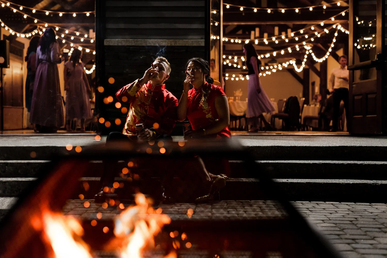 A couple dressed in traditional red and gold Asian attire sitting on steps by a fire pit, smoking and enjoying a night at a celebration or wedding reception with string lights and guests in the background.