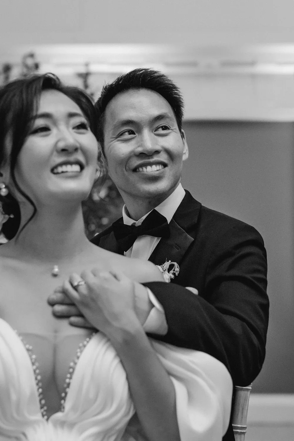 A black and white photo of a smiling bride and groom, the groom in a tuxedo with a bow tie, sitting closely together during their wedding celebration.