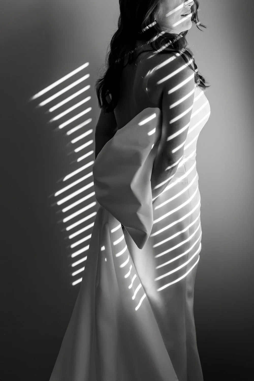 Black and white photo of a woman with wavy hair, standing sideways, partially covered by a large cloth or skirt, with shadows of horizontal blinds cast across her body.