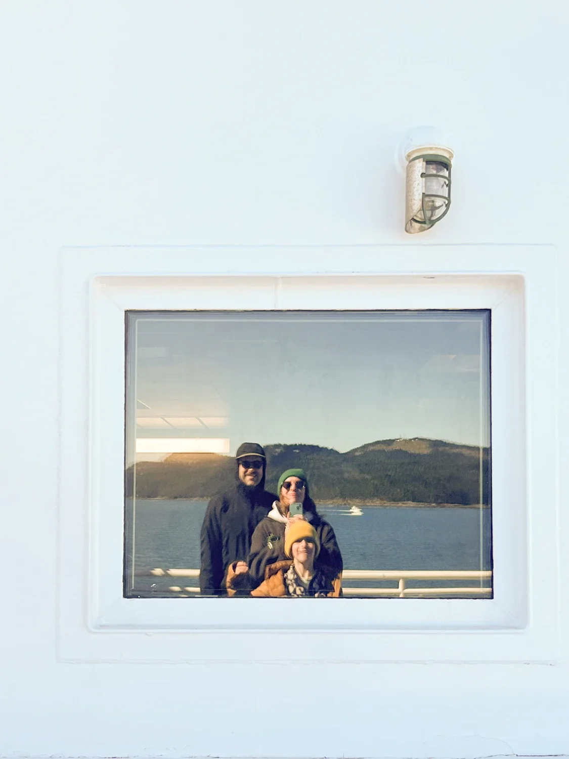 Three people taking a selfie in a window reflection on a boat, with water, mountains, and a boat in the background.