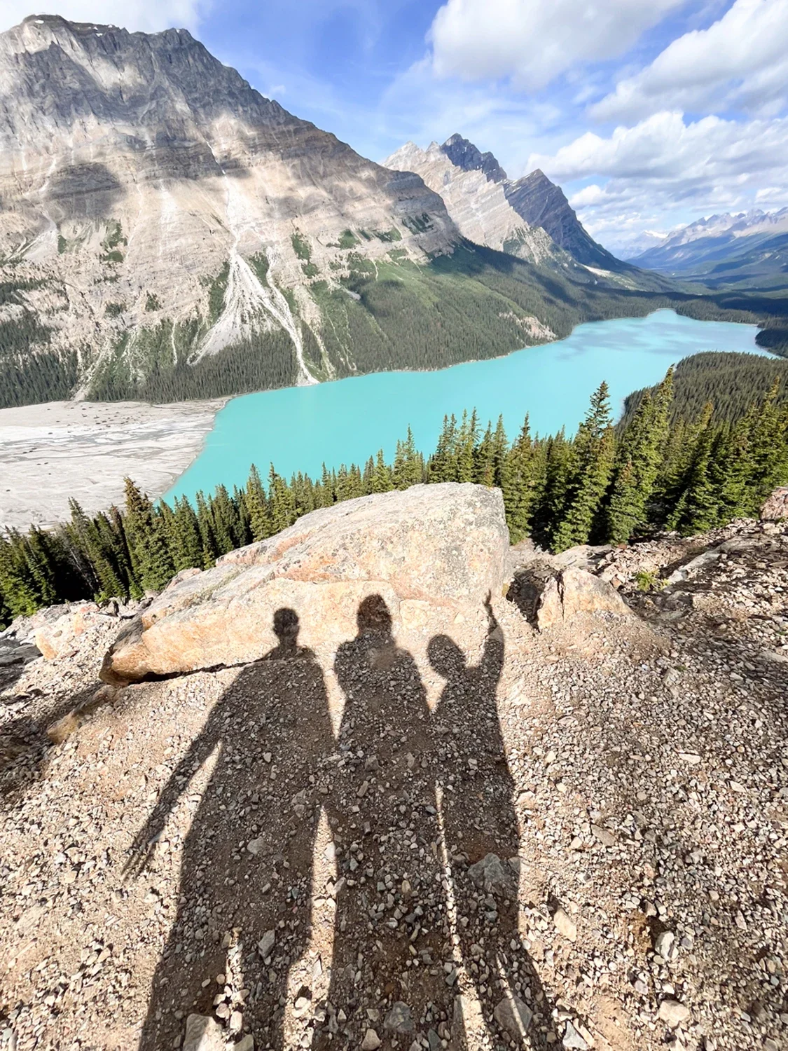 Shadows of three people cast on a rocky hillside overlooking a scenic mountain lake with a forested shoreline and rugged mountain peaks.