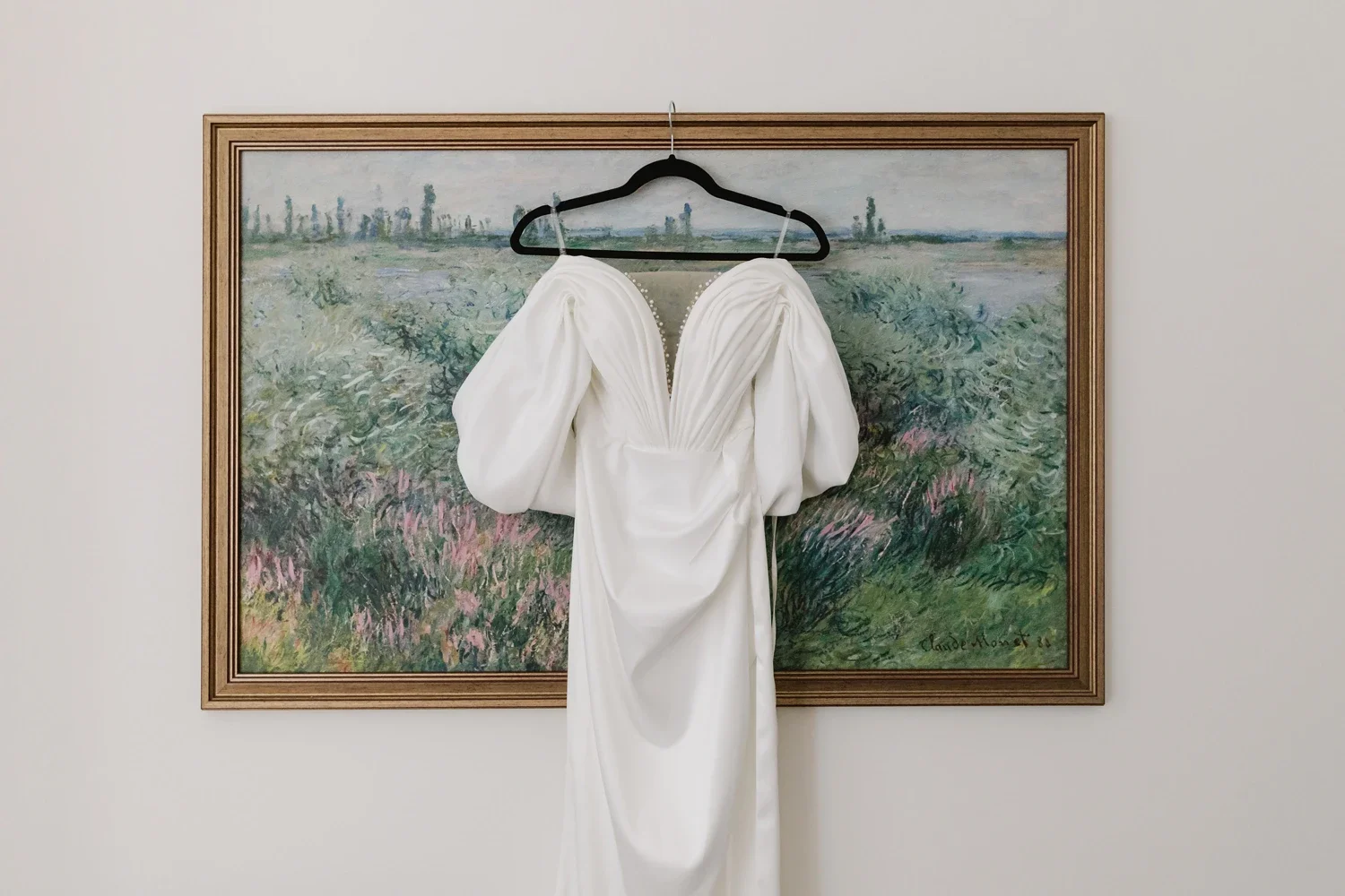 White wedding dress with puffed sleeves hanging on a black hanger in front of a framed landscape painting.