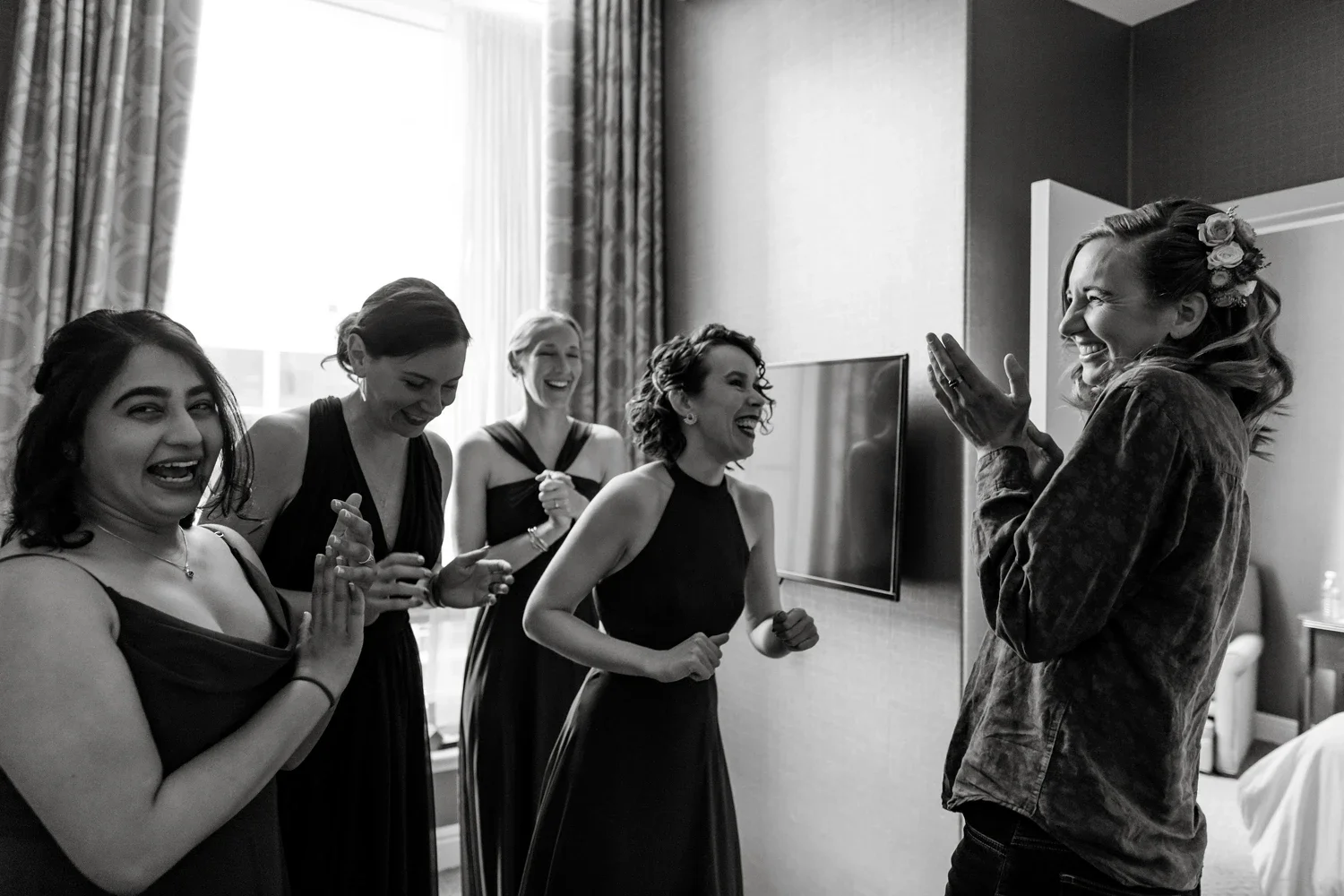 A group of women in a room, laughing and smiling, with one woman wearing flowers in her hair, while another woman is clapping and appears surprised or happy.