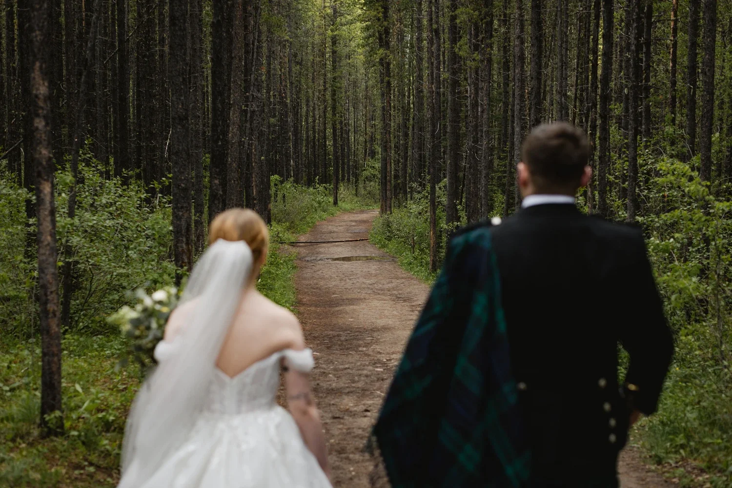A bride and groom walking on a dirt path through a green forest, seen from behind.