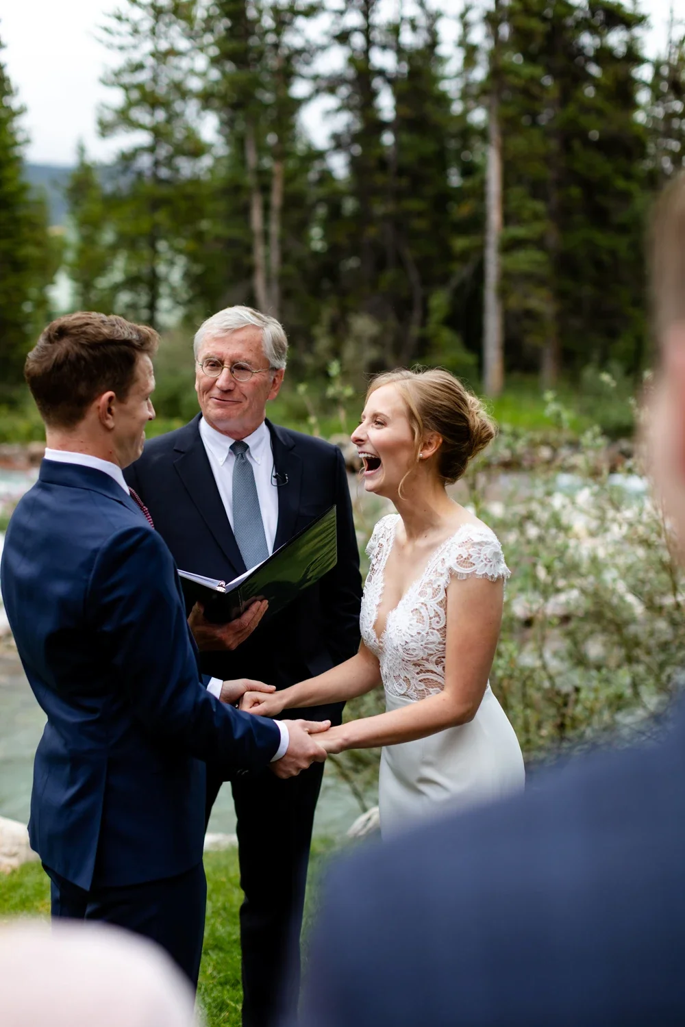 A wedding ceremony outdoors with a bride and groom holding hands, smiling, and an officiant reading from a book, surrounded by trees and nature.