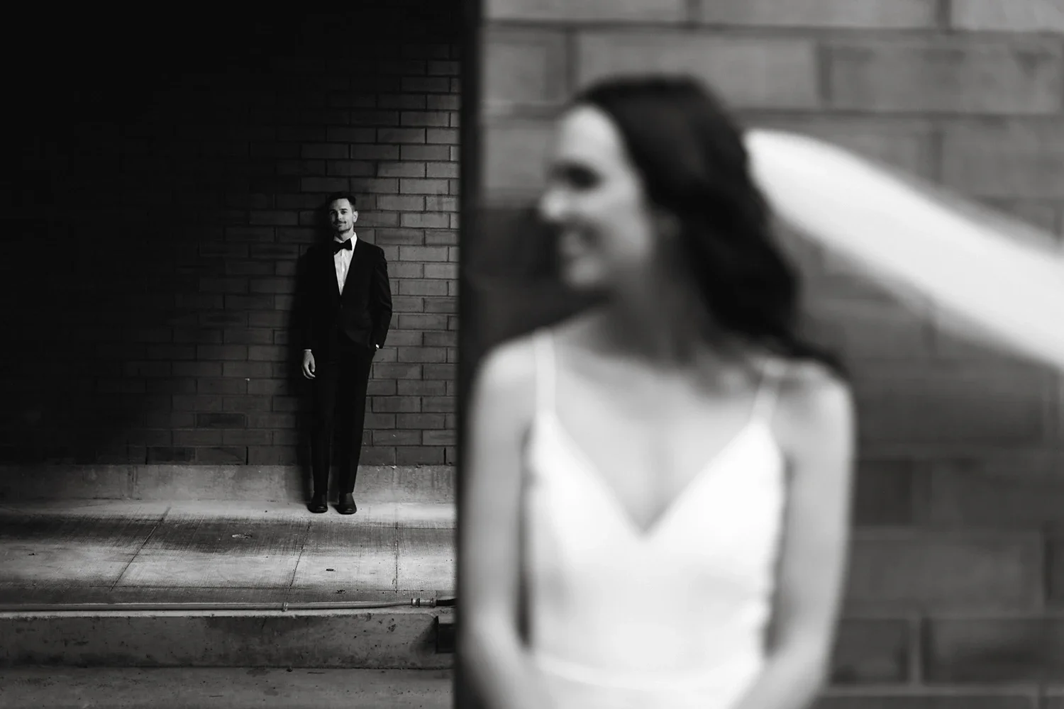 Black and white photo of a woman in a white dress with long dark hair, smiling and looking to the side, with a man in a tuxedo standing against a brick wall in the background.