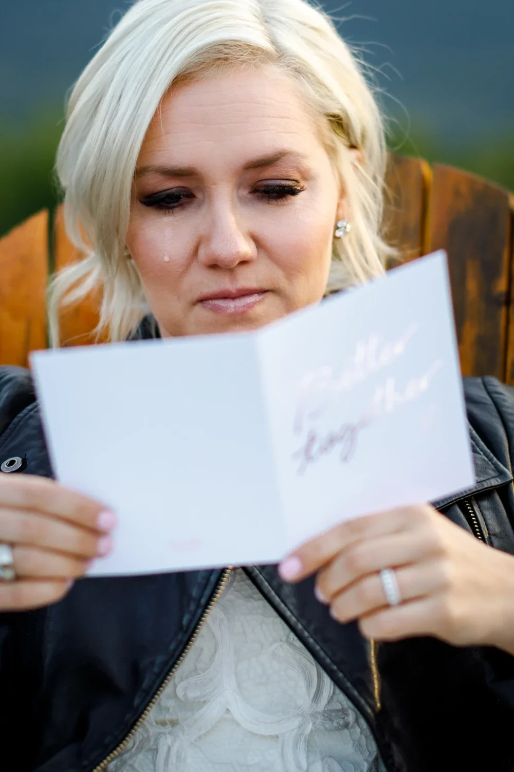 A woman with blonde hair and earrings is looking at a card she is holding, with a tear running down her face.