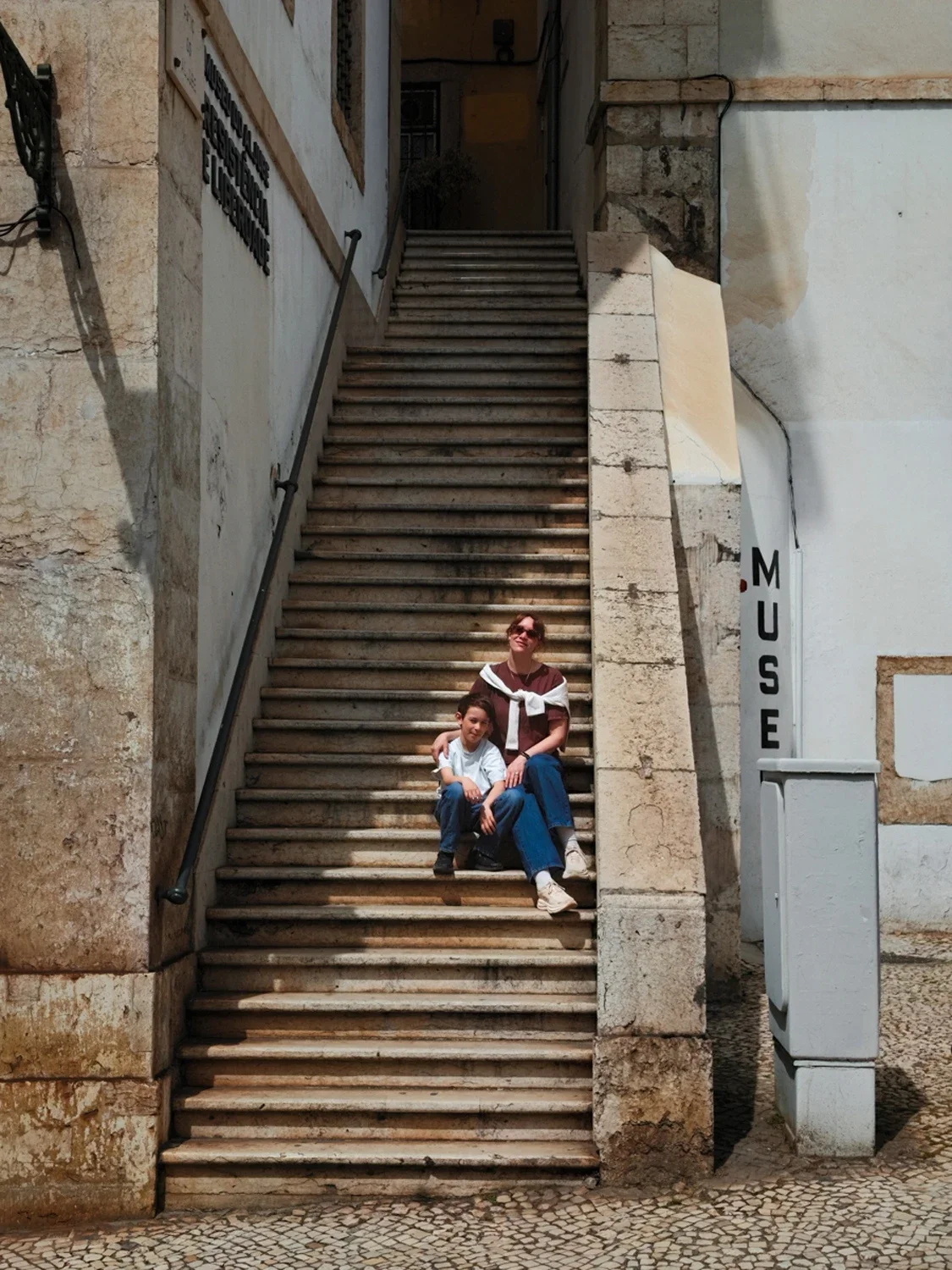 A woman and a young boy sitting on an outdoor stone staircase next to a museum sign. The woman has short reddish hair, sunglasses, and is wearing a t-shirt and jeans. The boy has dark hair and is wearing a white shirt and jeans.