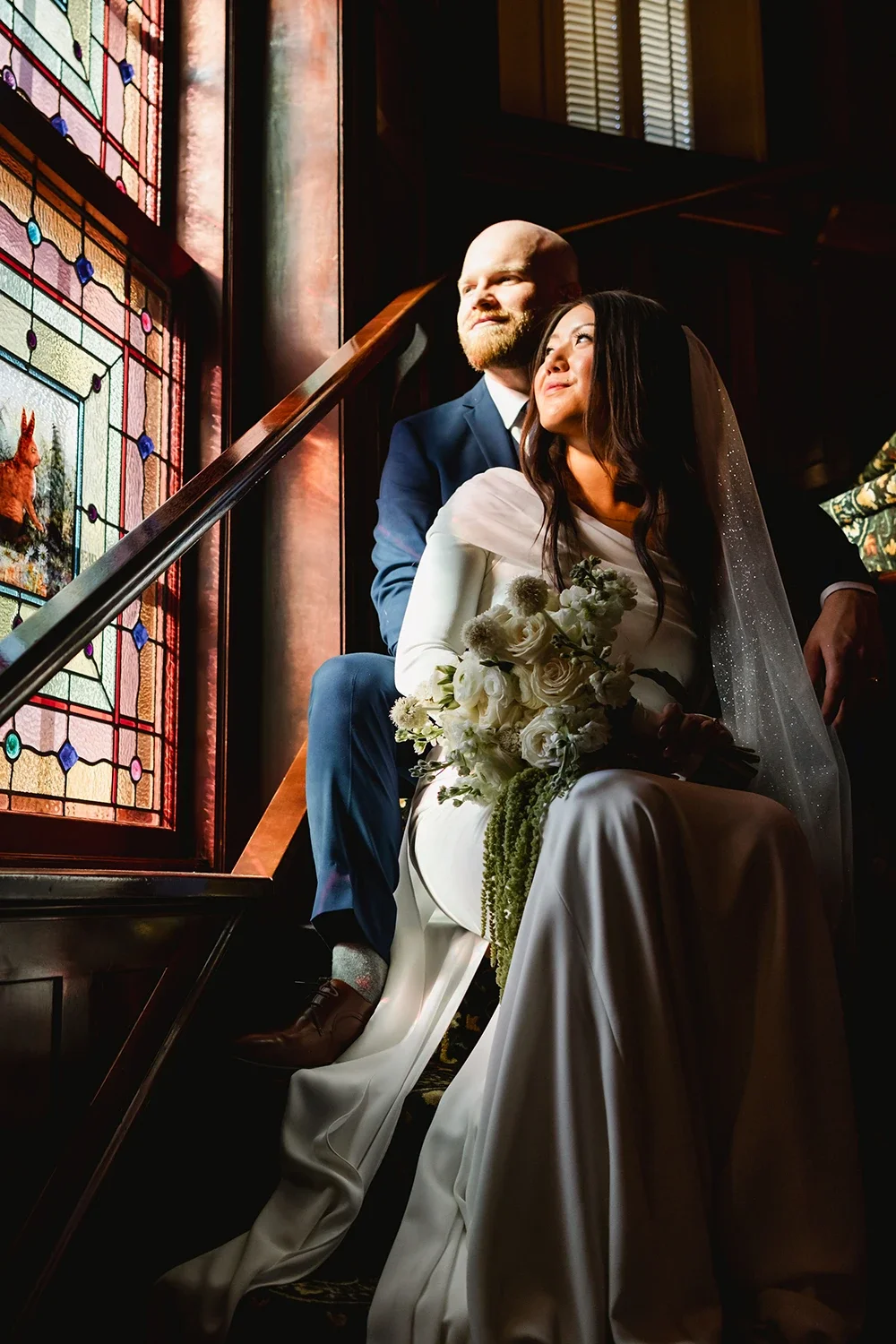 A newlywed couple sitting on a staircase inside a church, with stained glass windows behind them. The bride is holding a bouquet of white flowers, and both are gazing thoughtfully into the distance.