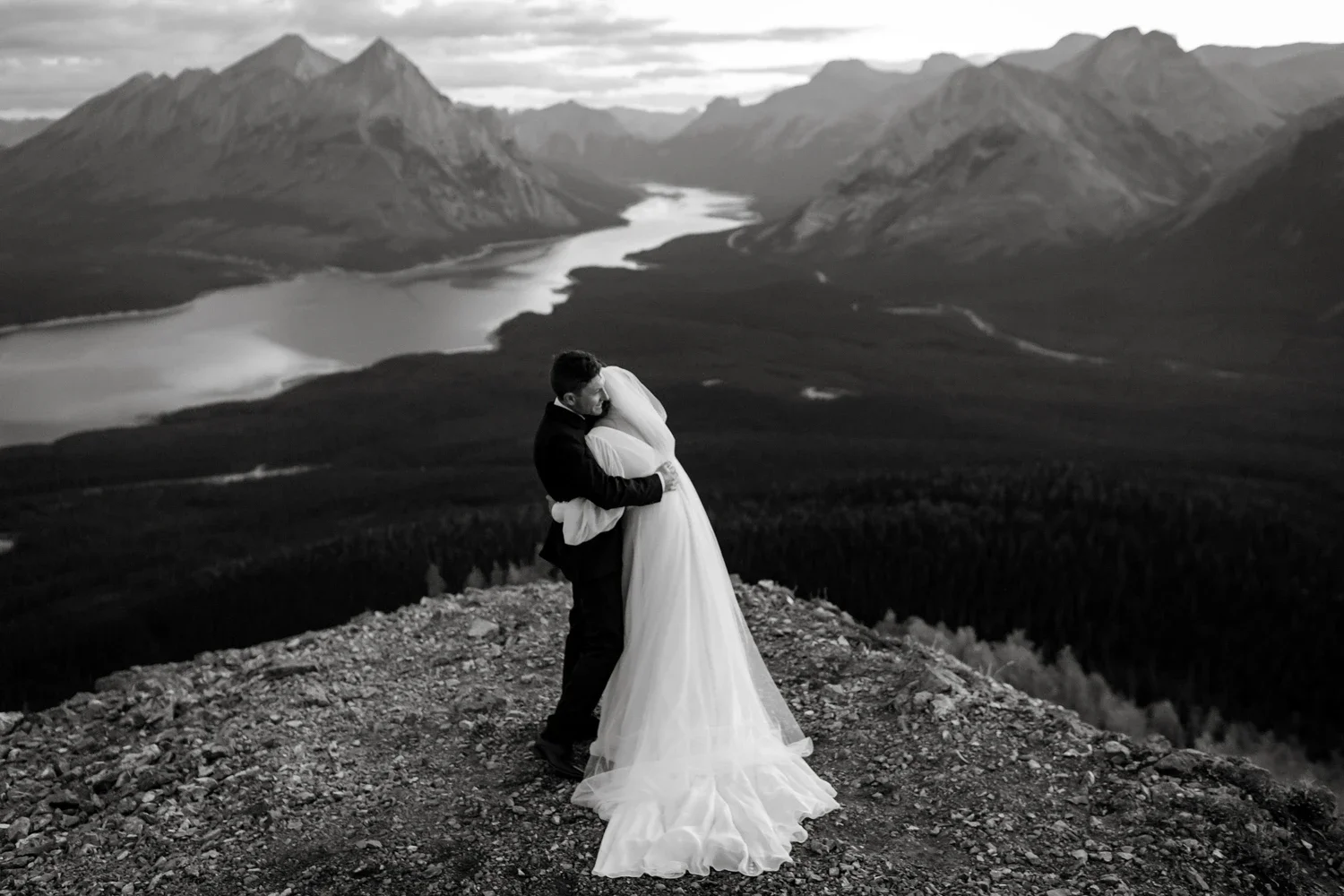 A black-and-white photograph of a couple hugging on a rocky mountain ledge with a scenic landscape of mountains, a lake, and forest in the background.