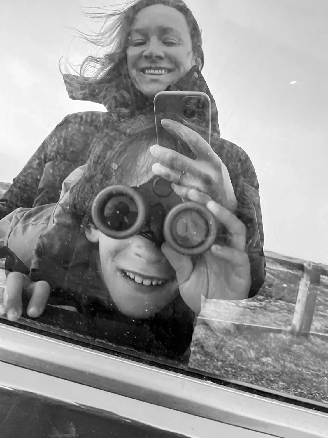 A woman and a child are taking a selfie in front of a reflective surface. The woman is smiling, holding a smartphone, and has windblown hair. The child is smiling and holding a pair of binoculars.