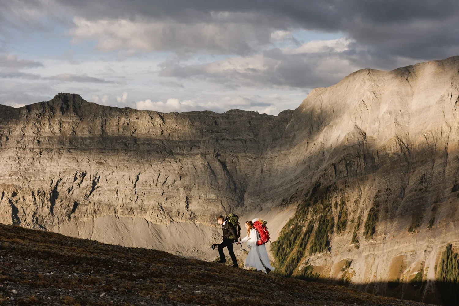 A couple hiking in a mountainous area with rocky cliffs and sparse vegetation during daytime.