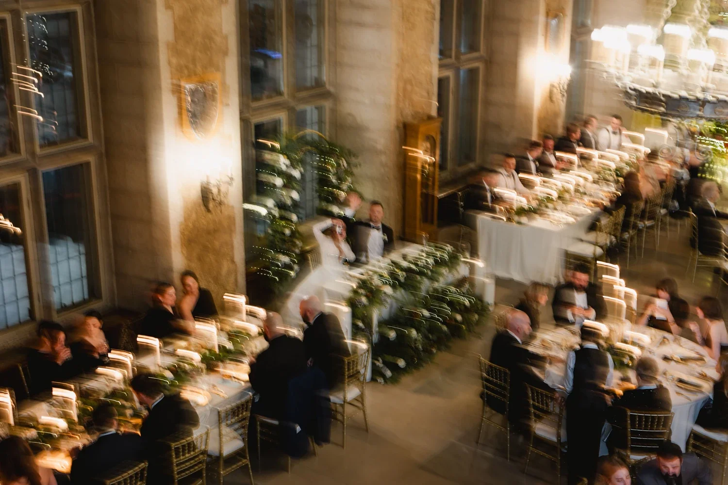 A blurry photo of a formal dinner event in an elegant hall with high ceilings, large windows, and decorative wall features. Guests are seated at multiple long tables, dressed in tuxedos and evening gowns.
