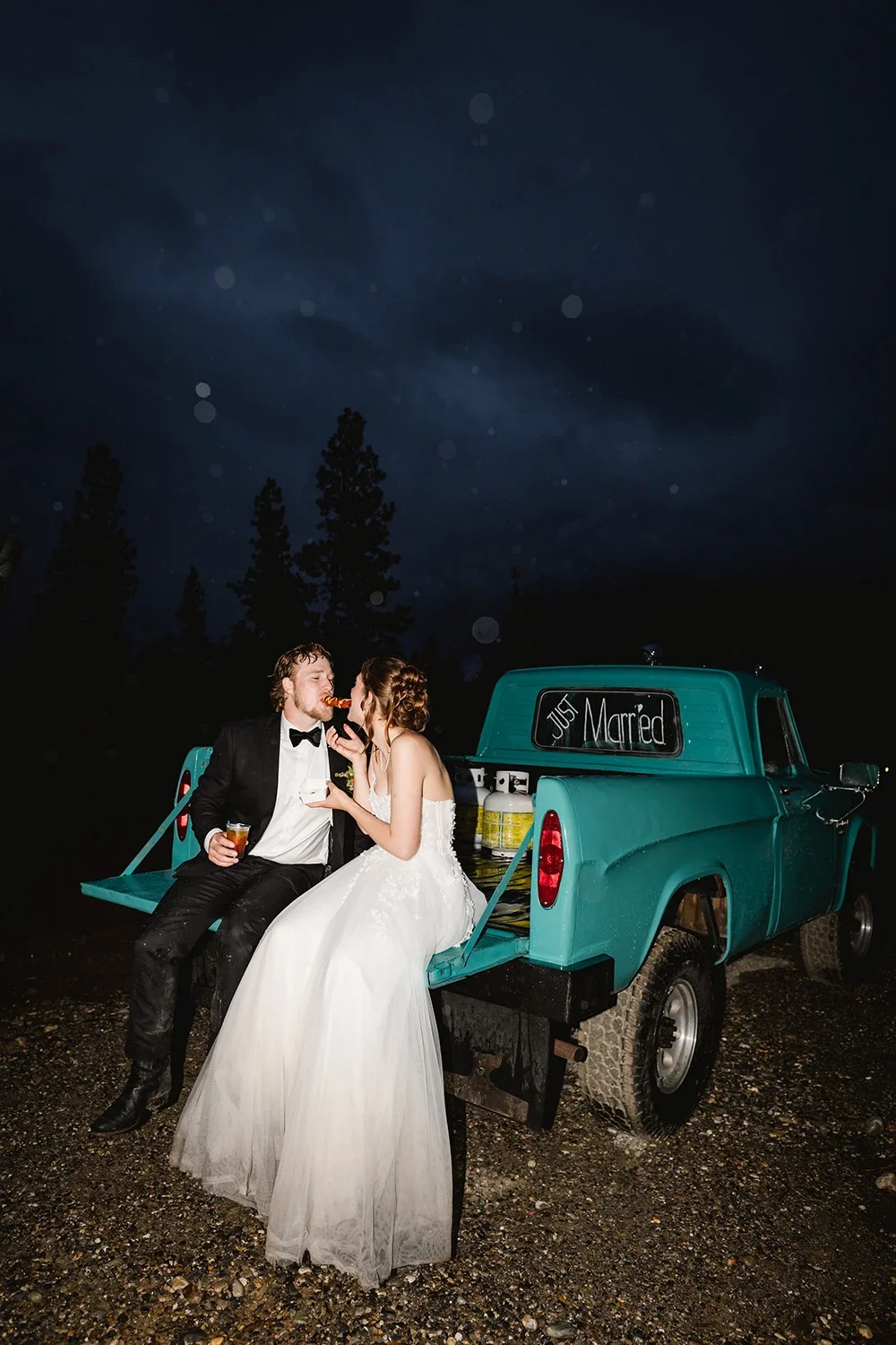 A newlywed couple in wedding attire sitting on the back of a vintage turquoise pickup truck, sharing a piece of pizza and drinks at night. The truck has a sign on the window that reads 'Just Married', and there are propane tanks in the truck bed. The