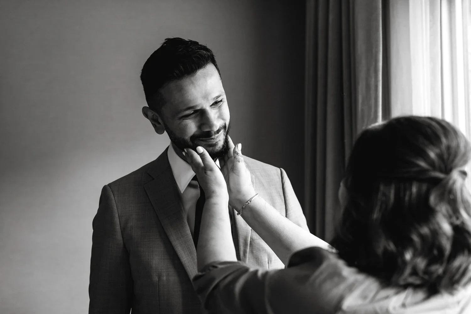 A woman adjusts a groom's face while he smiles, wearing a suit, in a softly lit room with curtains.