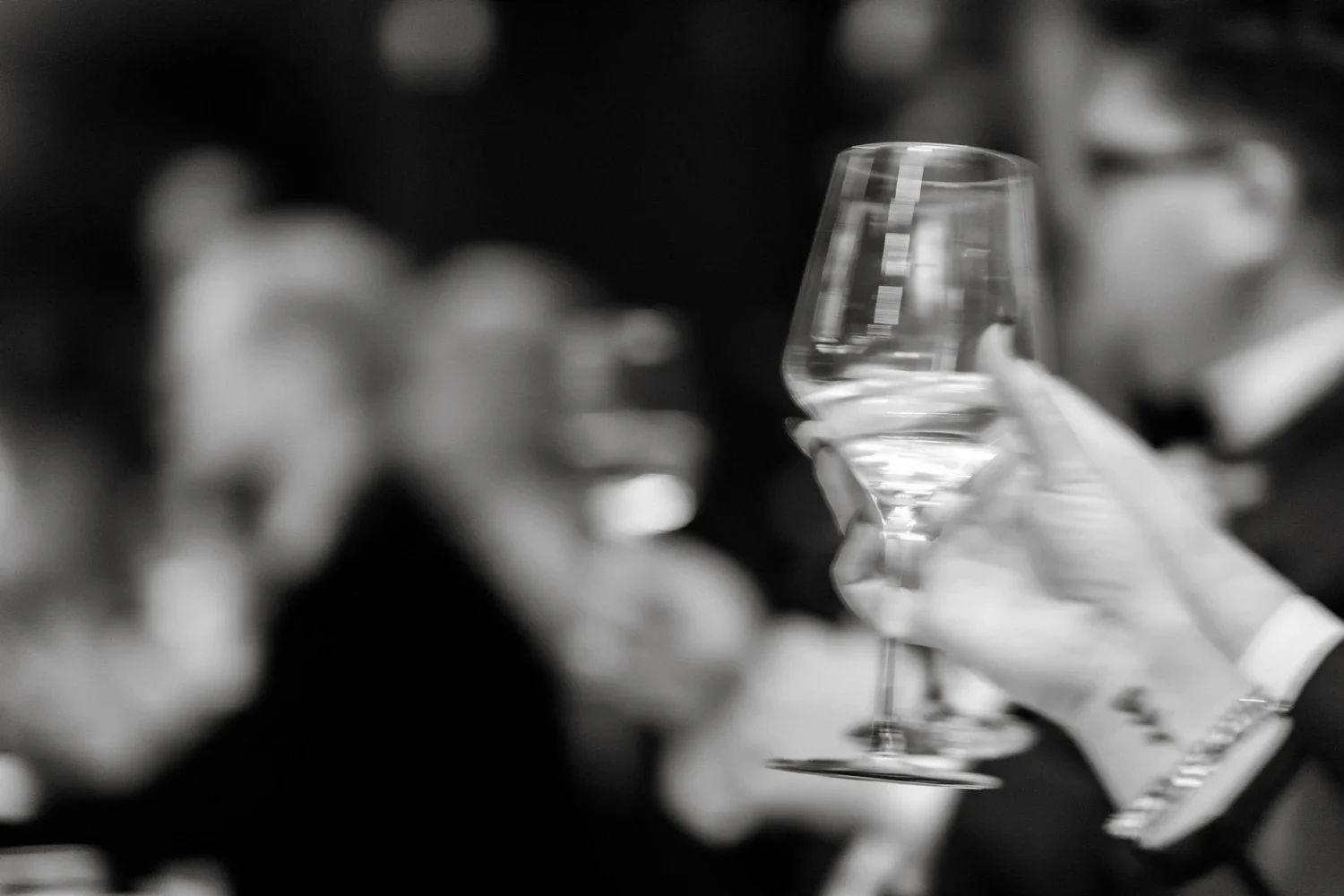 A person holding a wine glass filled with water, with other glasses and people blurred in the background.