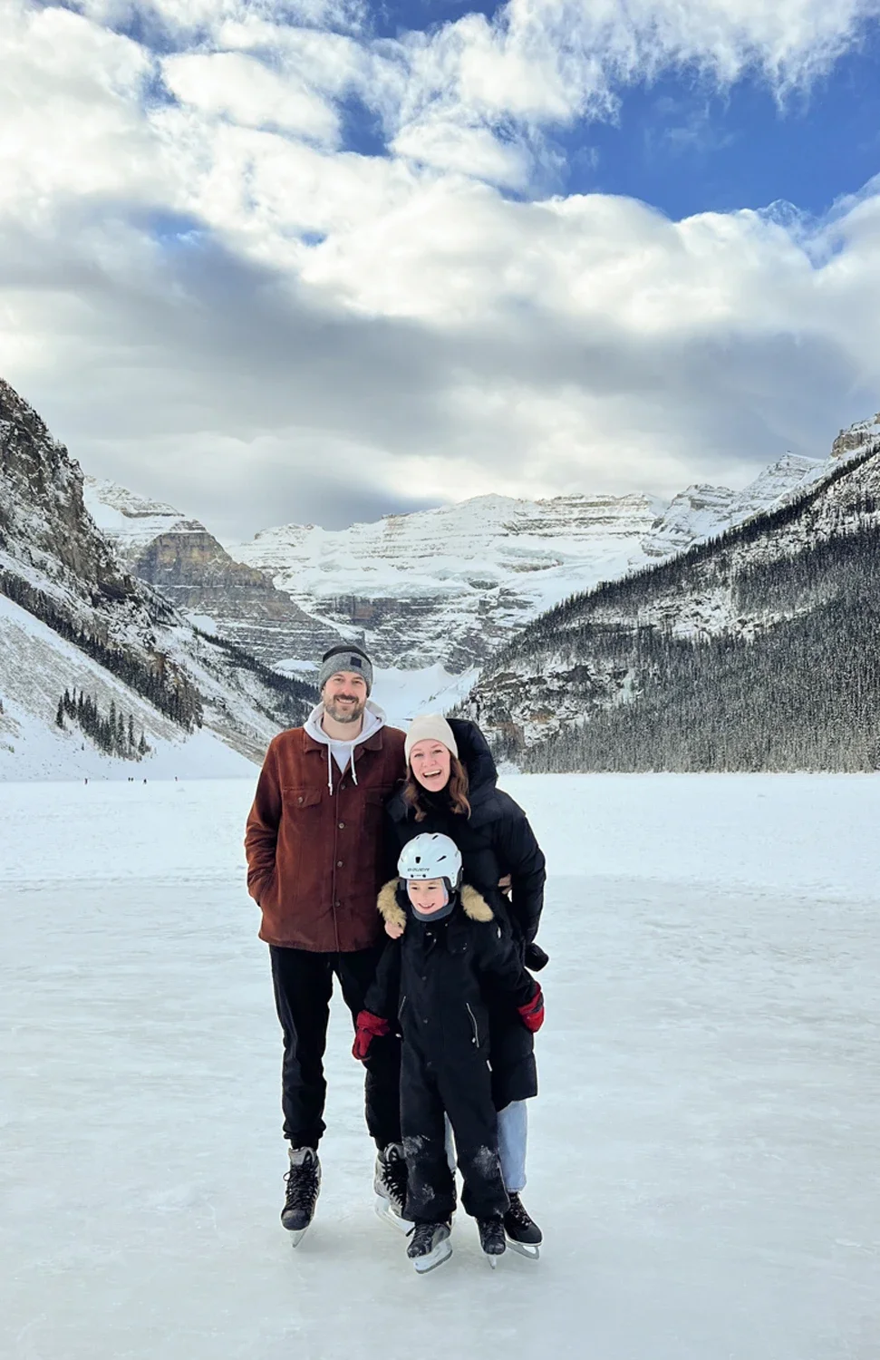 Family of three ice skating on a frozen lake with snow-covered mountains and trees in the background.