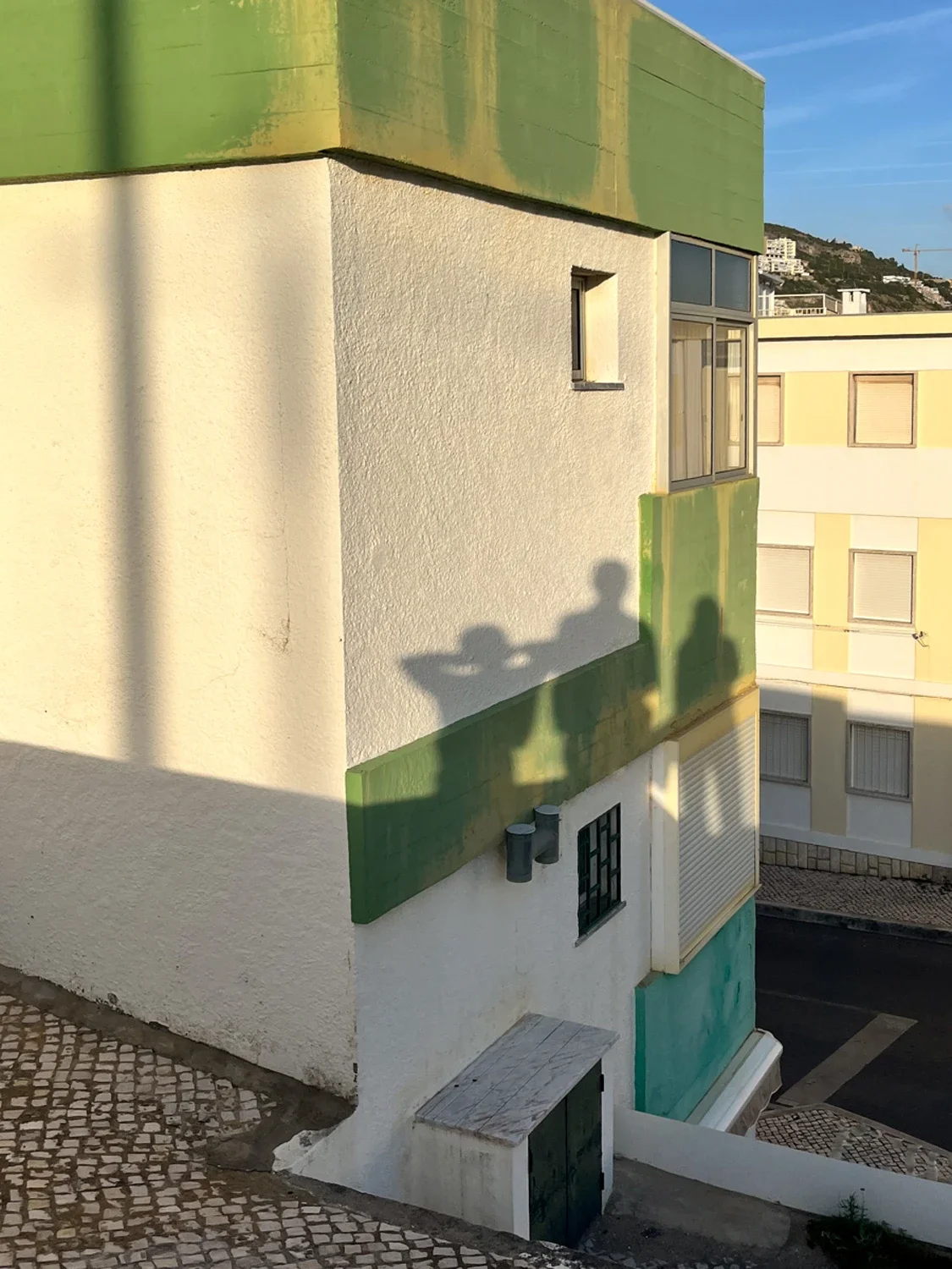 View of a corner of a colorful residential building with white, green, and yellow exterior walls, and a small window. Shadows of people are cast on the wall, and there are other buildings and a hillside in the background.