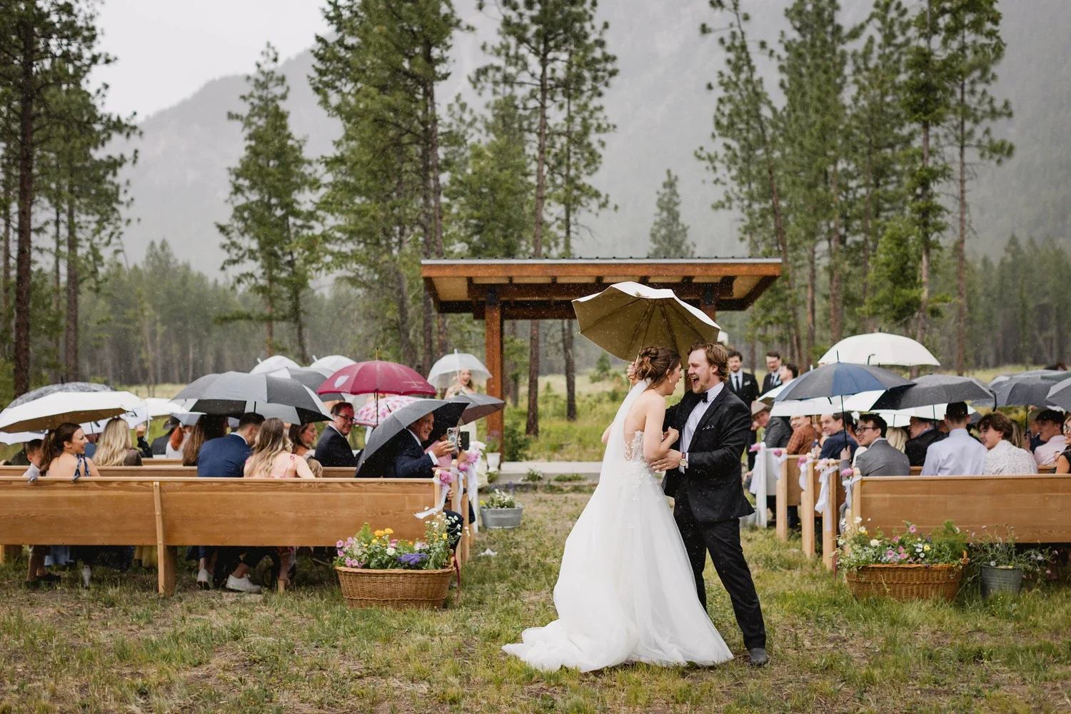Bride and groom dancing under a brown umbrella during an outdoor wedding in the rain, with guests seated in benches holding umbrellas in a forested area.