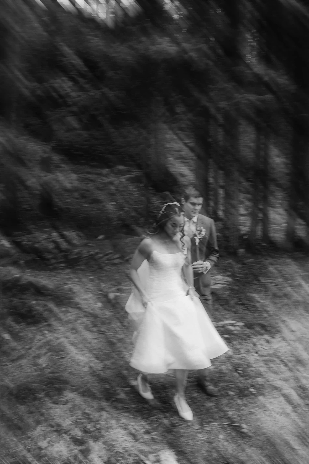 A black-and-white photograph of a bride and groom walking outdoors on a dirt path surrounded by trees.