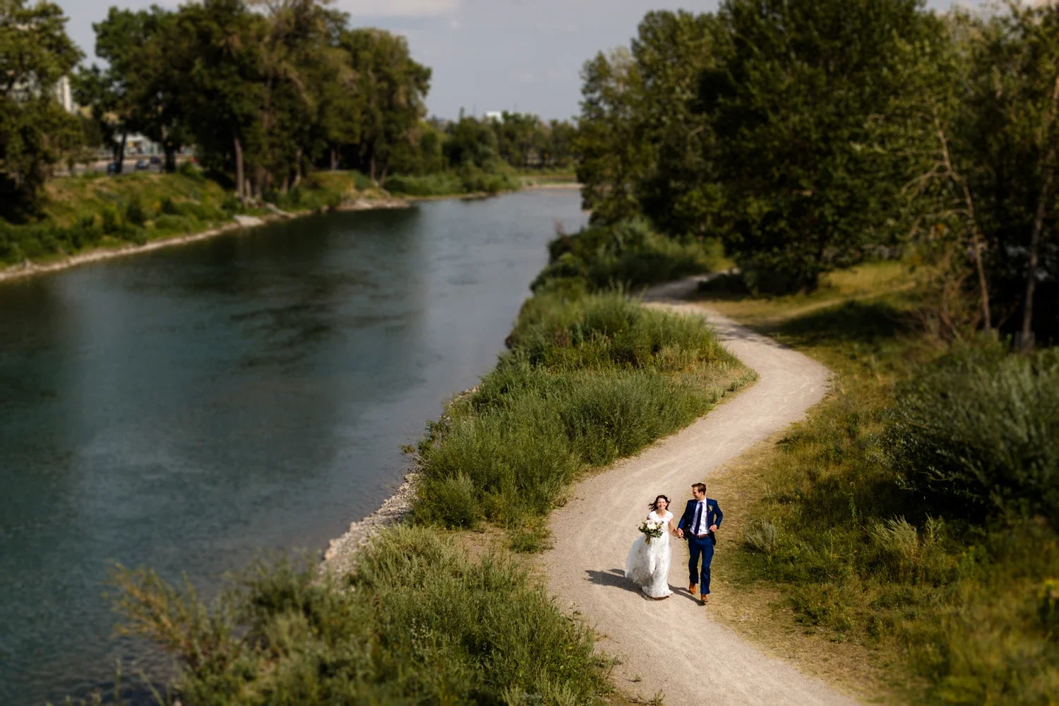 A bride and groom walking hand in hand along a winding dirt path next to a river surrounded by trees and greenery.