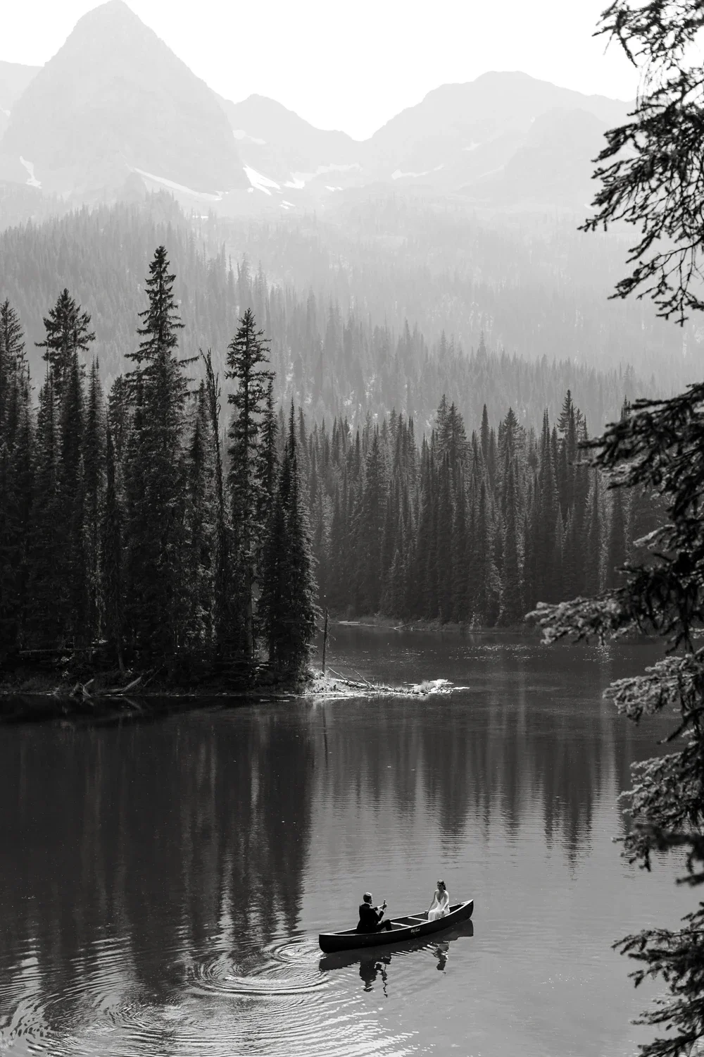 A couple in a boat on a calm river surrounded by dense forested mountains in black and white.