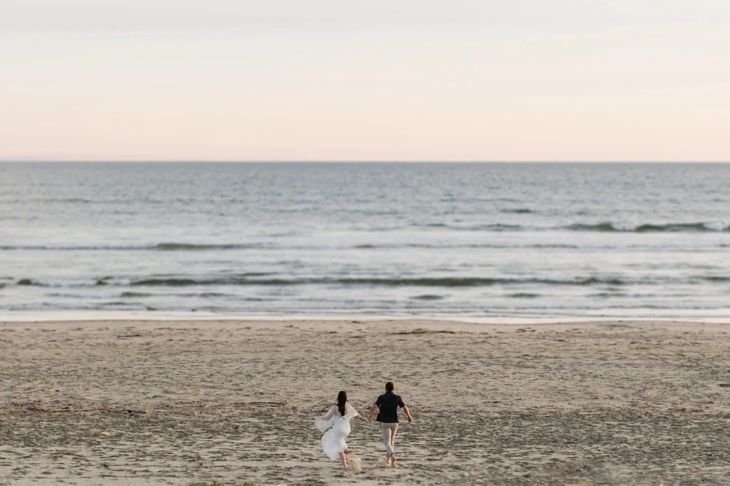 A couple dressed in white and dark clothing is running on a sandy beach towards the ocean.