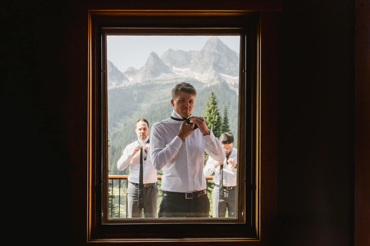 Group of four men adjusting their ties outside a window with a mountainous landscape in the background.
