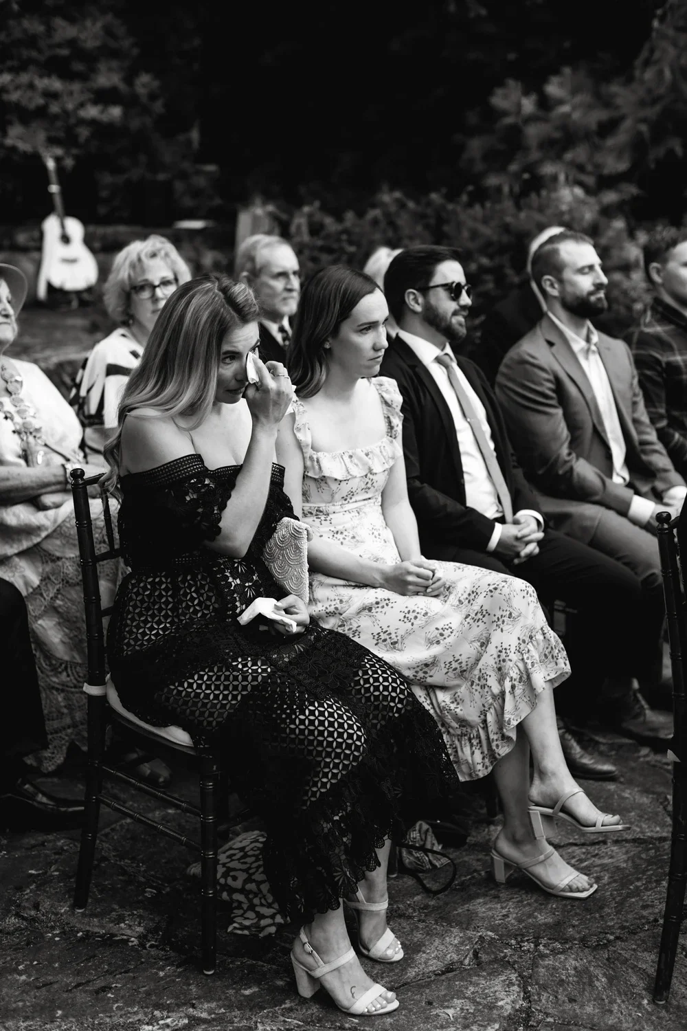 Group of people sitting outdoors, attending a formal event, with one woman wiping away a tear.