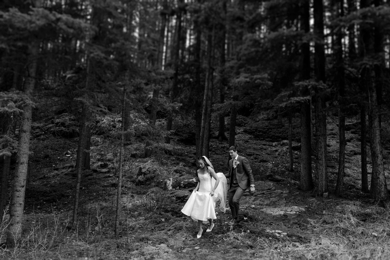 A black and white photo of a bride and groom walking through a forest. The bride is wearing a white wedding dress and veil, and the groom is in a suit. They are holding hands as they walk on a wooded trail.