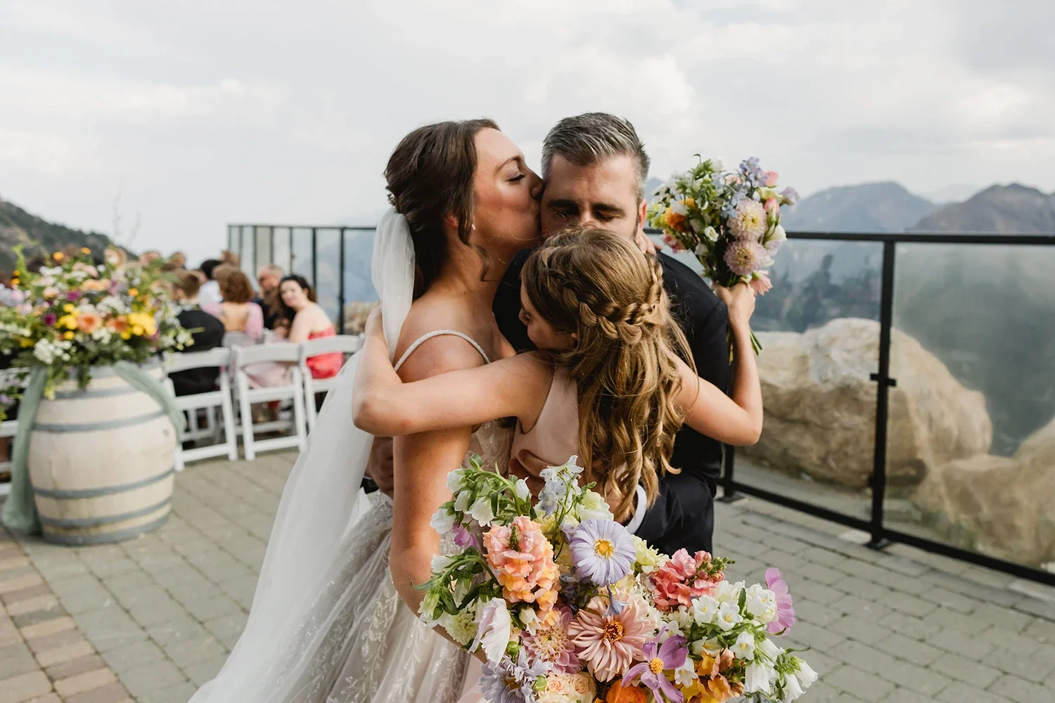 A bride, groom, and a young girl, likely a flower girl, hugging each other with happy expressions at a wedding reception. The bride is wearing a white wedding dress, and the young girl has long, wavy hair with braids. The bouquet contains various colorful flowers. In the background, there are guests seated at tables with floral arrangements, an outdoor setting with mountains, rocks, and a cloudy sky.