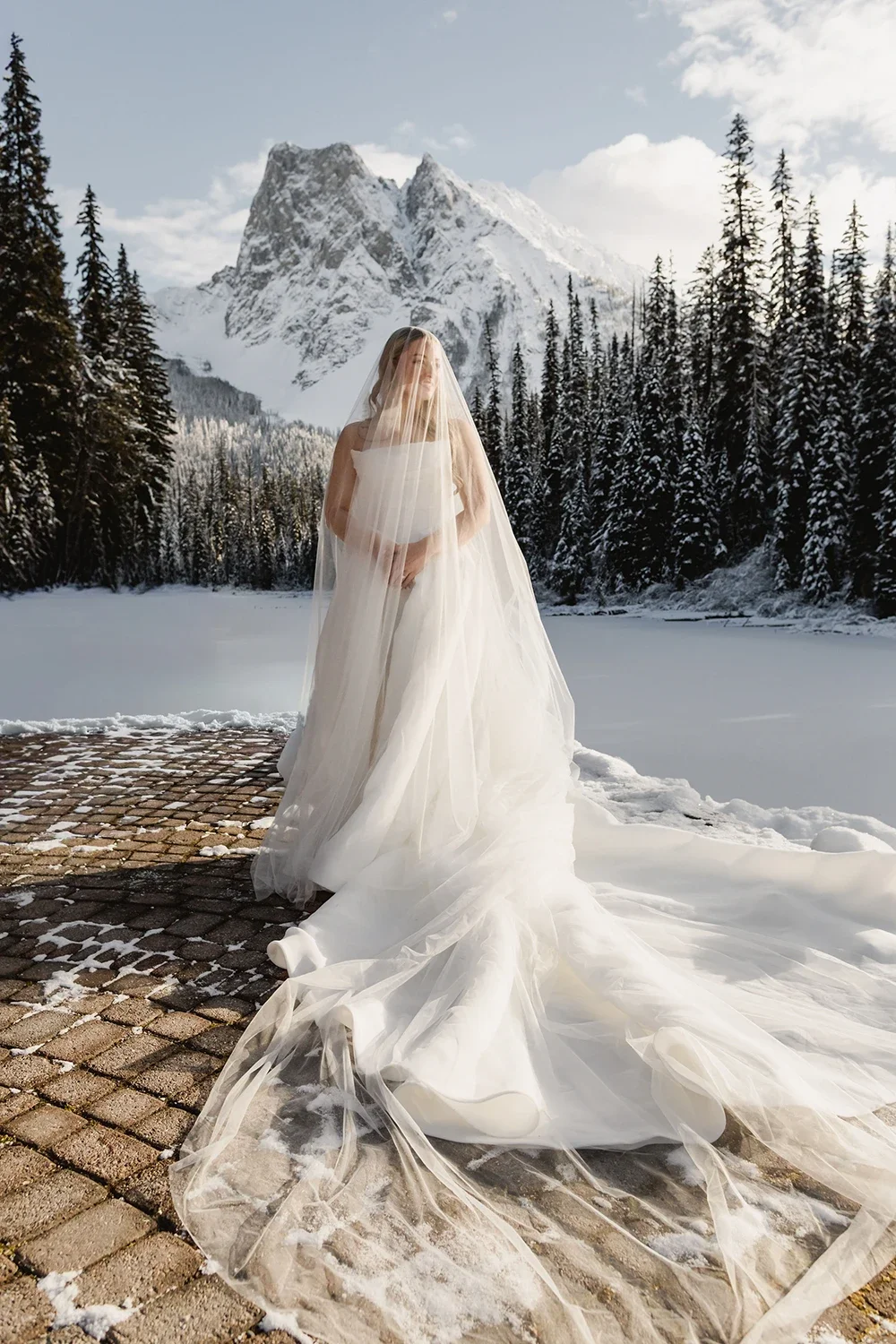 A bride in a wedding dress standing outdoors on a snowy brick pathway, with a snow-covered lake, tall evergreen trees, and a mountain in the background.