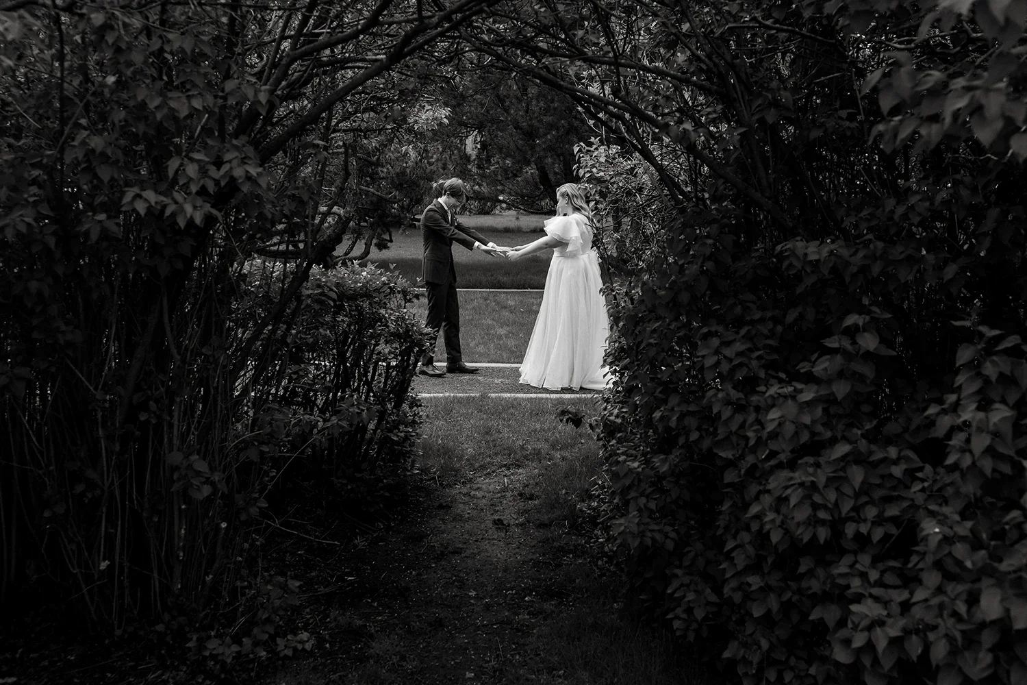 Black and white photo of a wedding couple holding hands in a garden, framed by bushes and trees.