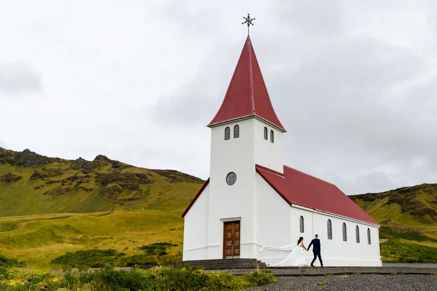 A bride and groom holding hands outside a white church with a tall red steeple on a cloudy day with green hills in the background.