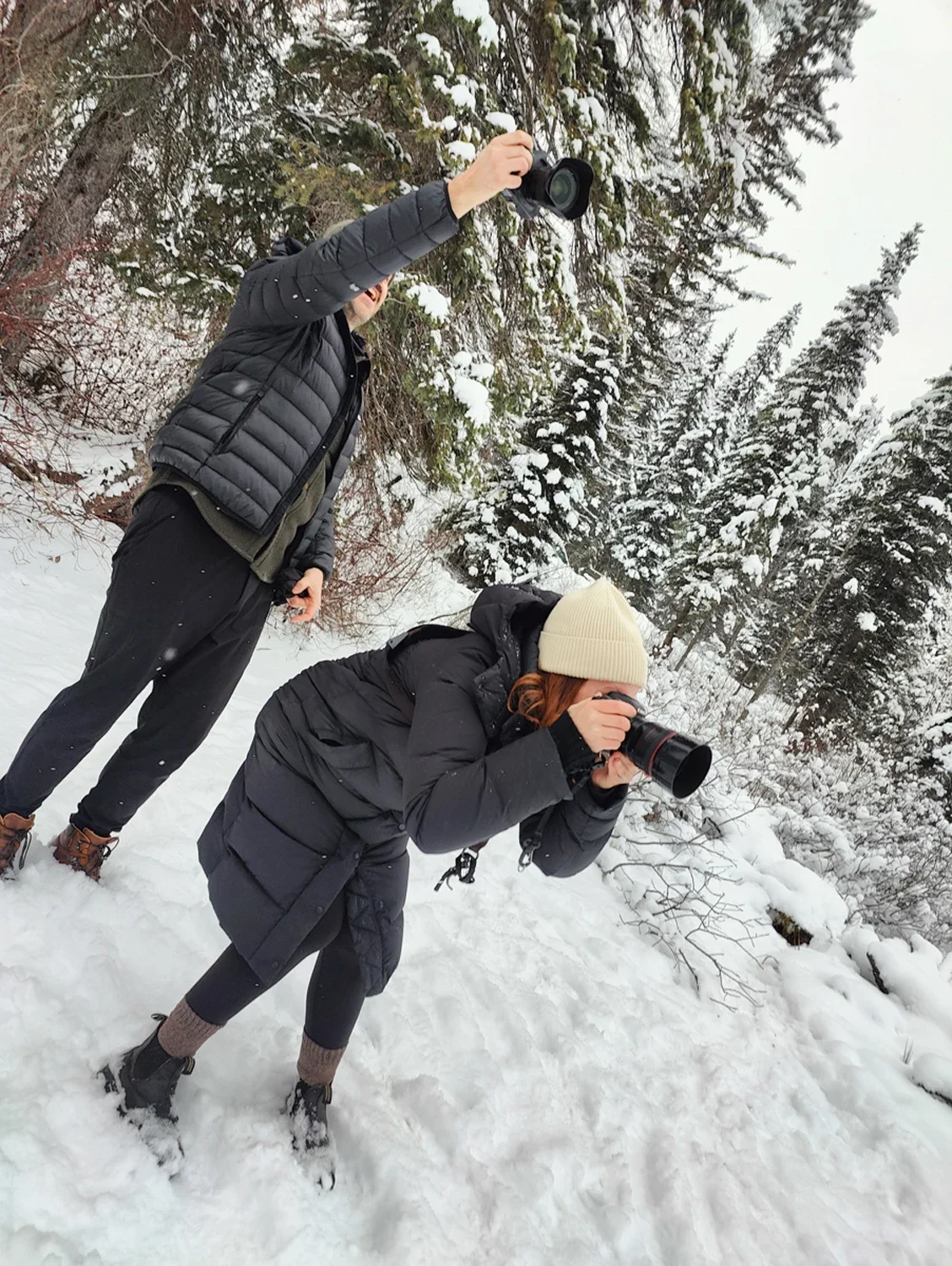 Two people in winter clothes, one woman and one man, taking photos with cameras in a snowy, forested area with snow-covered trees.