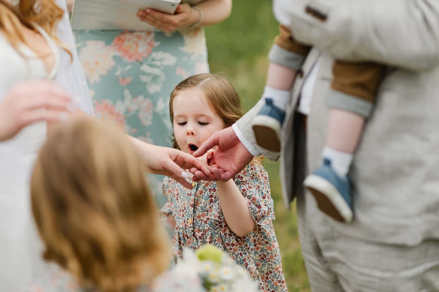 A young girl at an outdoor gathering touches the hand of an adult, with other children and adults nearby.