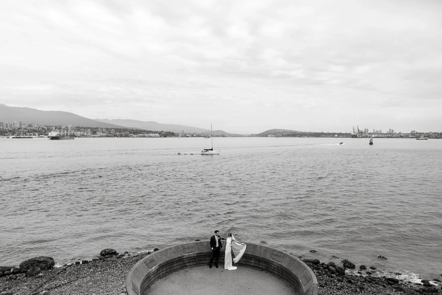 A black and white photo of a couple in wedding attire standing on a circular stone structure by the water, with boats and a cityscape across the river in the background.