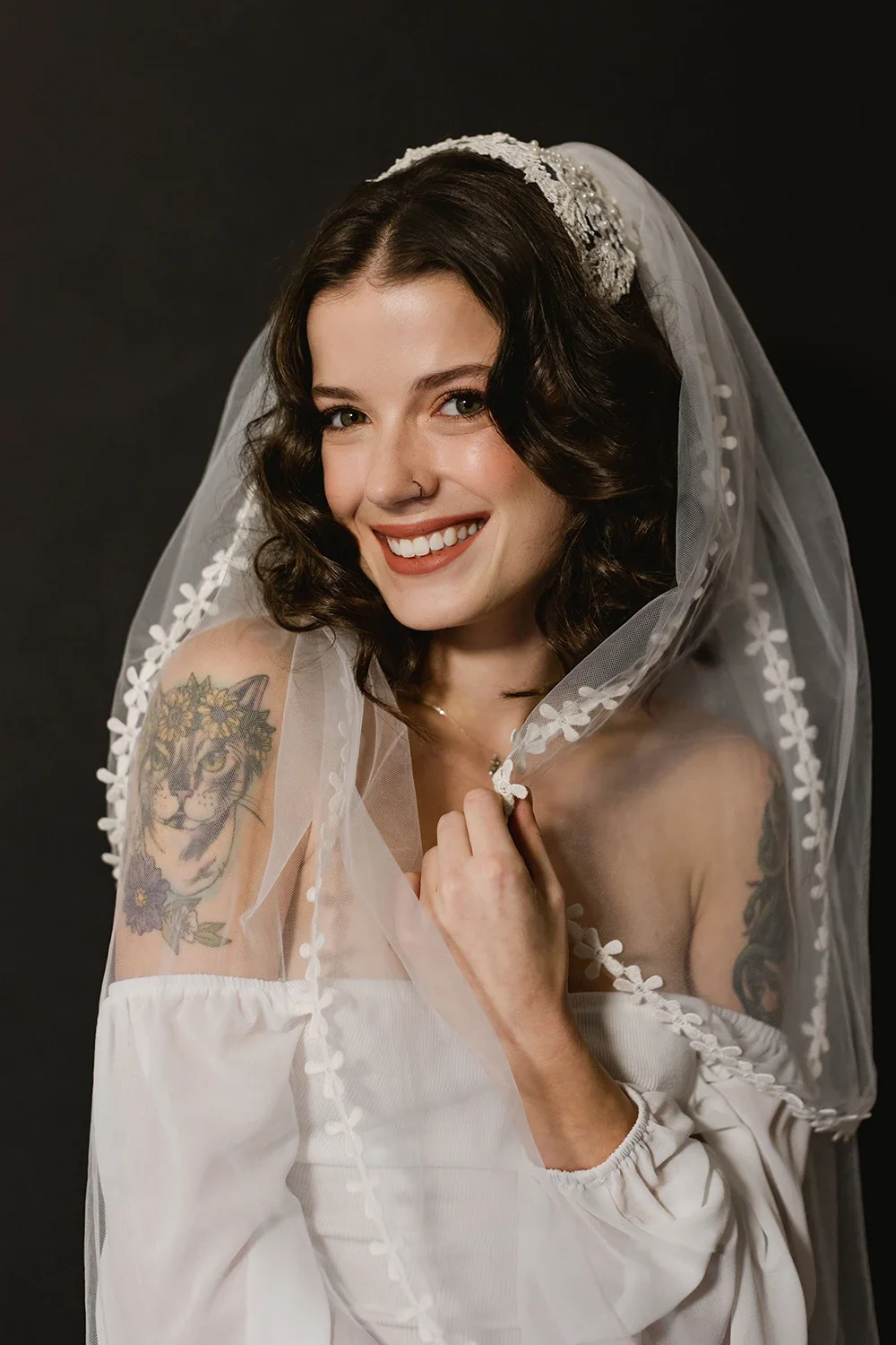 A young woman with dark wavy hair and a nose piercing smiling, wearing a white off-shoulder dress and a lace veil. She has a colorful tattoo of a lion and flowers on her left shoulder and is holding the edge of her veil.